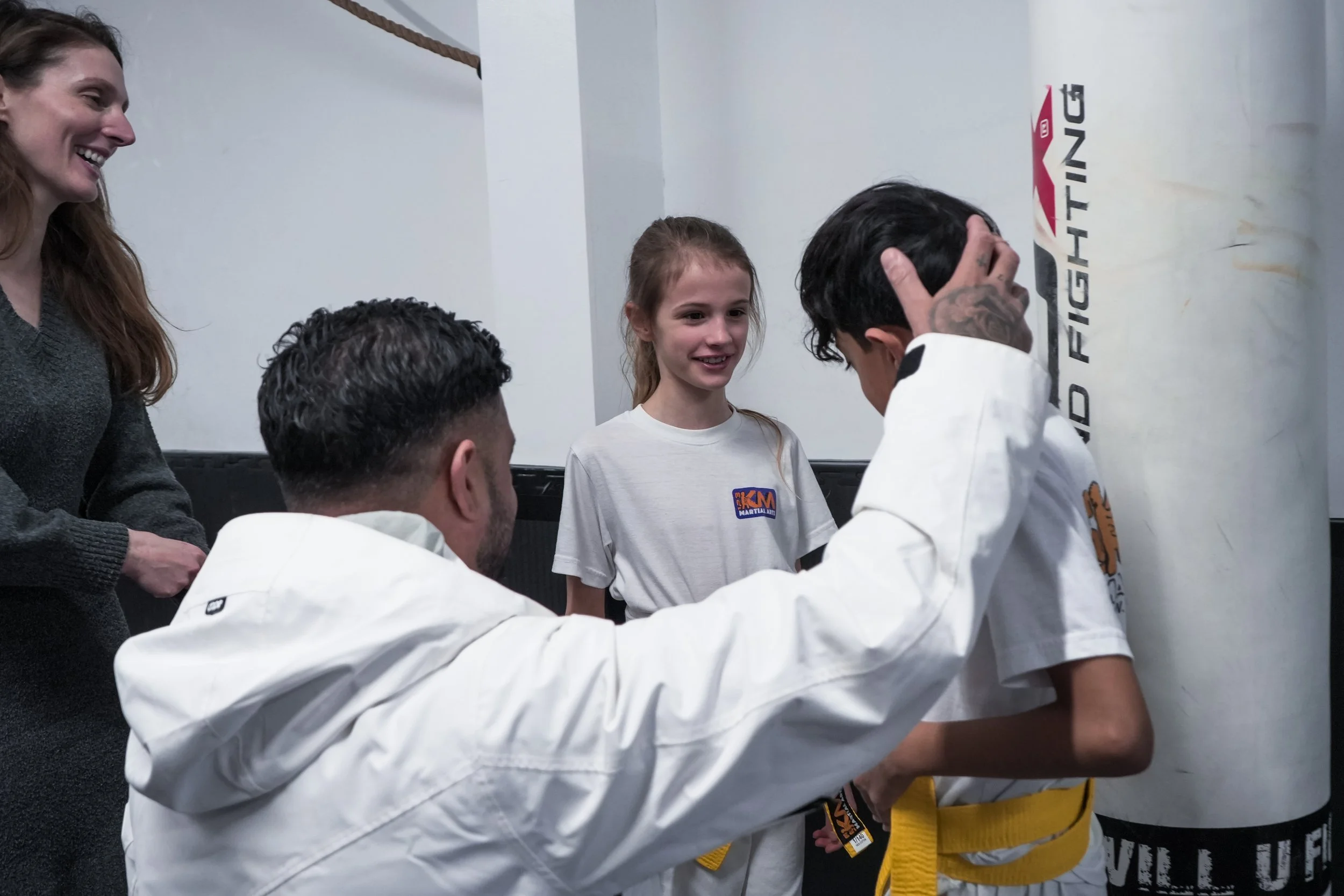 Children and adults in martial arts uniforms in a gym, with two kids in yellow belts and a girl in a white uniform with a patch, talking and smiling.