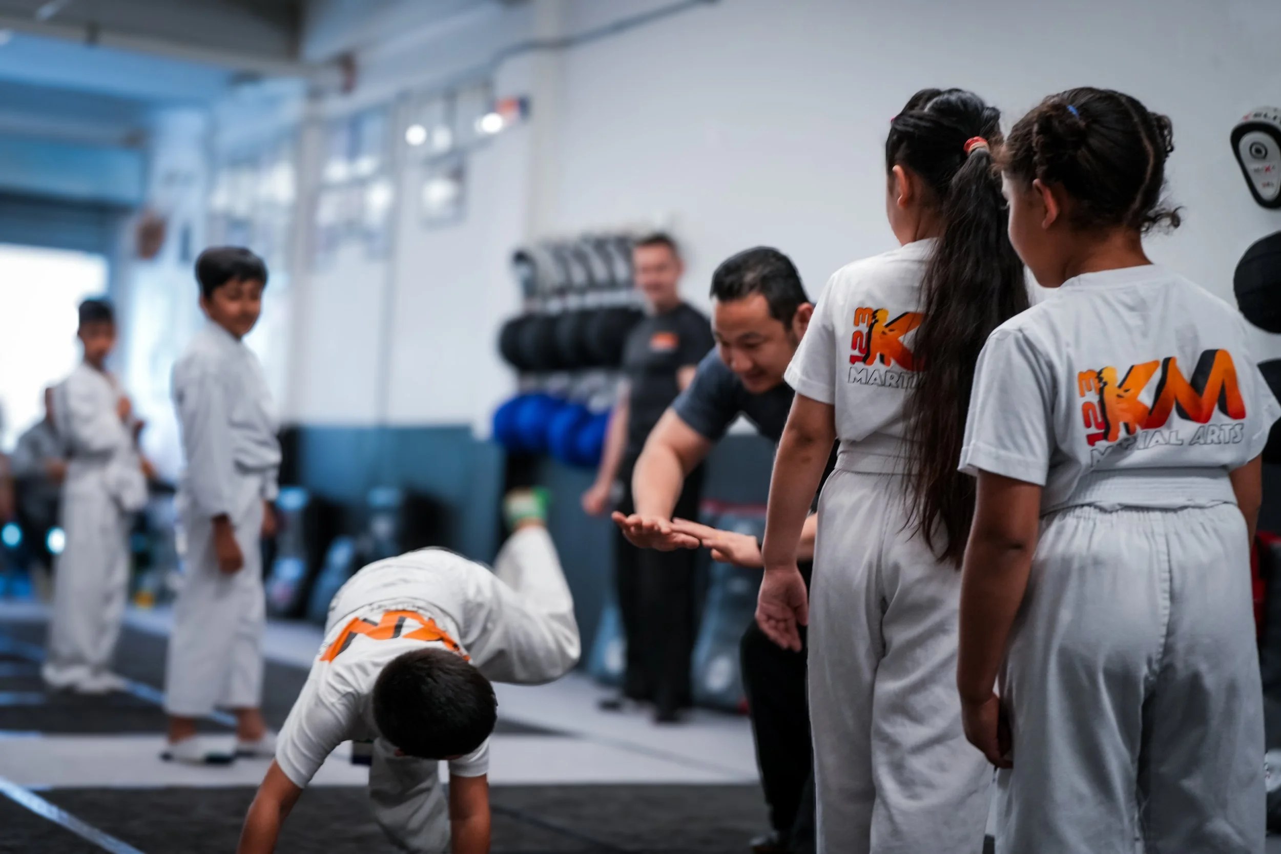 Children in martial arts uniforms practicing in a dojo, with a sensei instructing in the background.