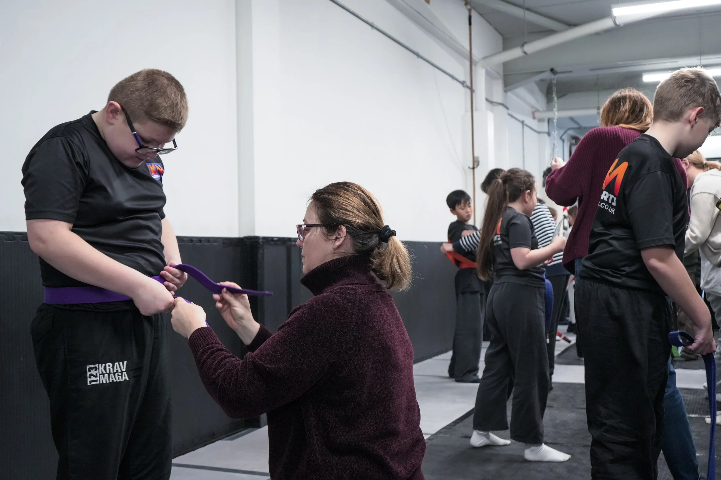 A young boy receiving a purple martial arts belt from a woman during an award ceremony in a gym. Several other children and adults are lined up in the background.