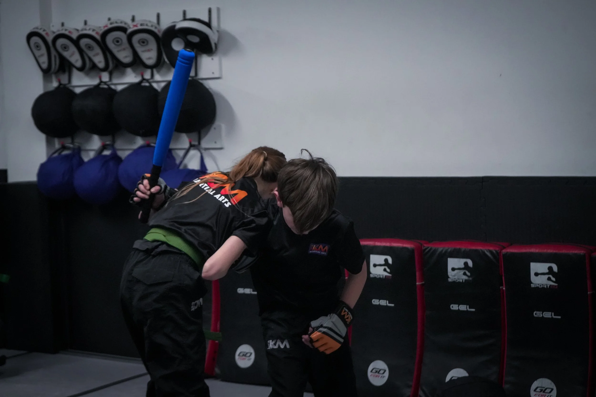 Two children practicing martial arts in a gym, with martial arts equipment and padded ring visible in the background.