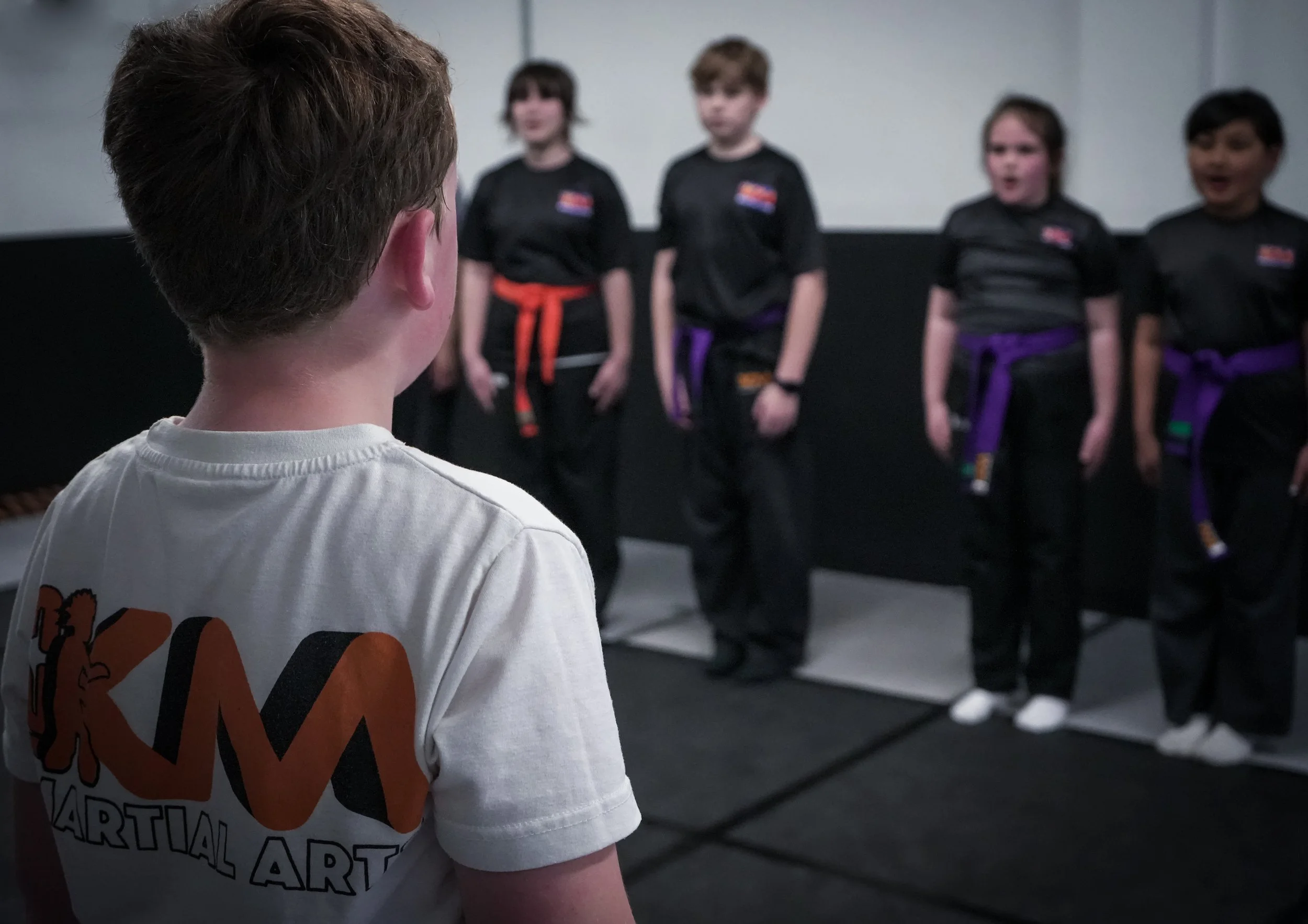 Young boy in a white martial arts uniform with hands together in a praying position, standing against a gray concrete-textured background.
