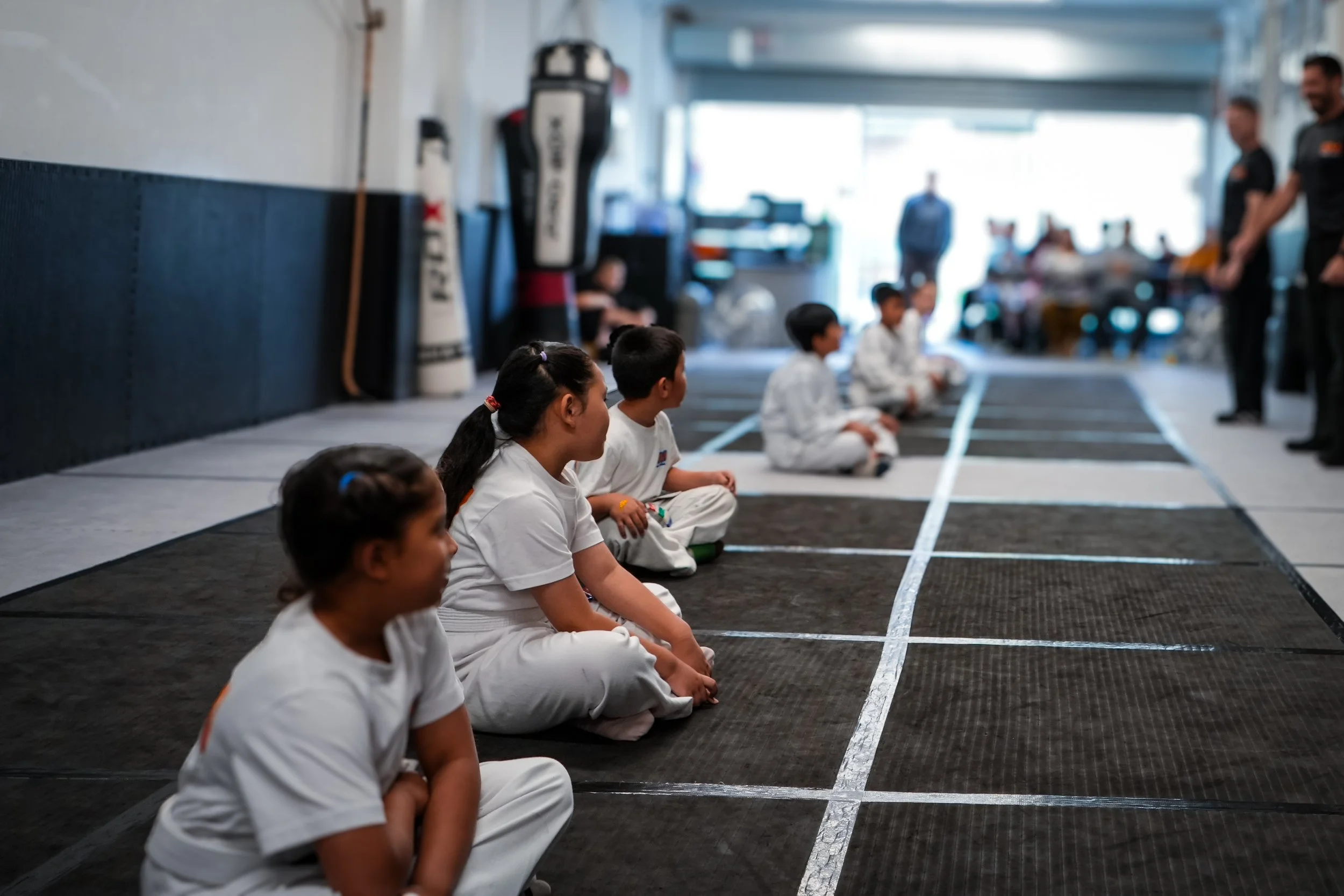 Children sitting cross-legged on the floor in a martial arts training facility, listening to an instructor, with training equipment such as punching bags in the background.