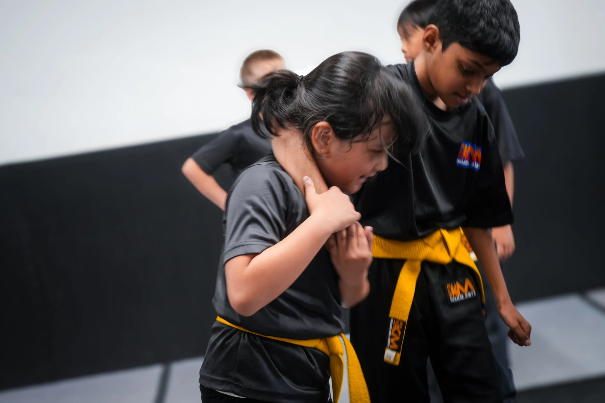 Two children wearing martial arts uniforms and yellow belts are practicing martial arts together, with one child assisting the other in a training move inside a gym.