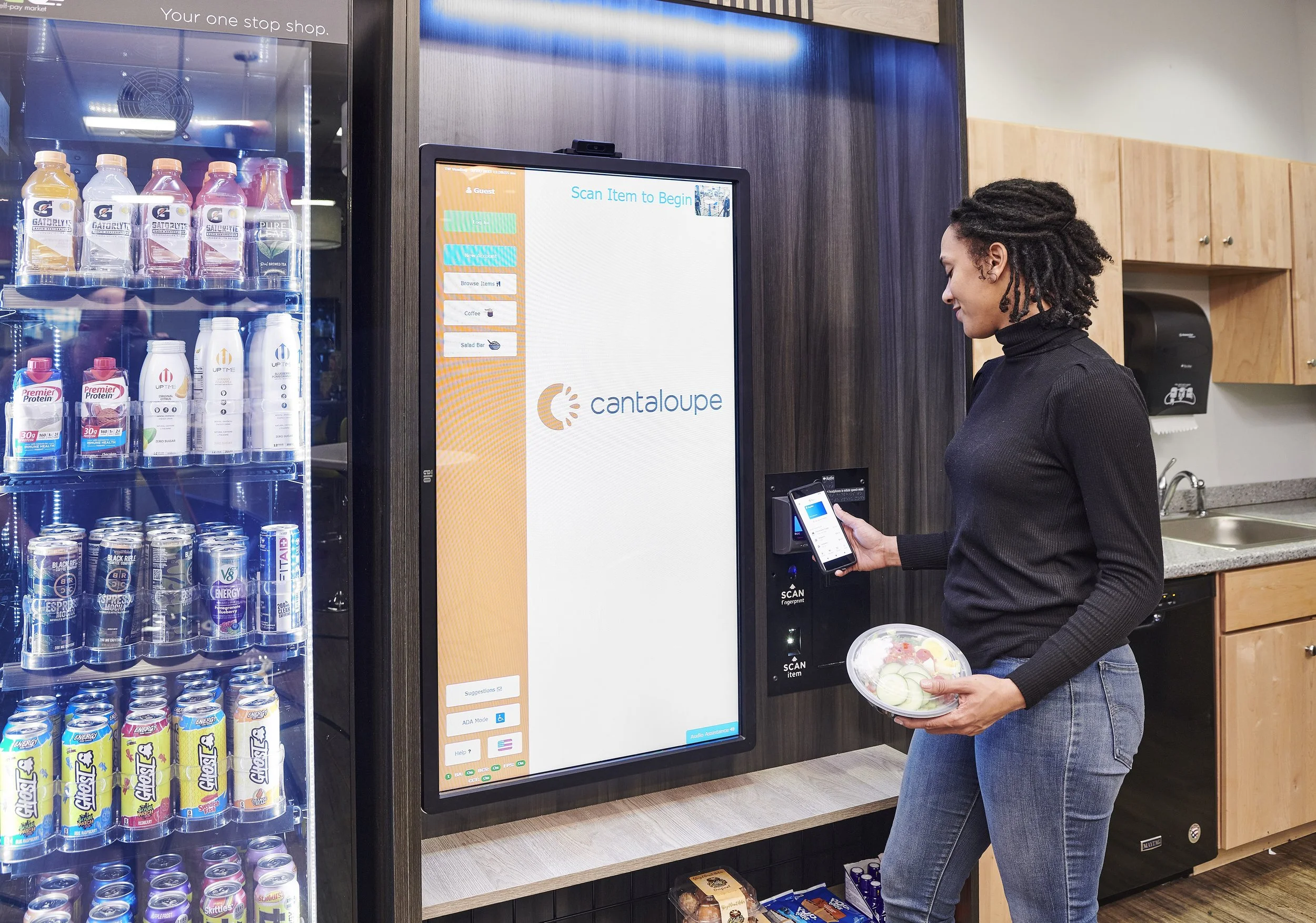 A woman using a mobile phone to scan an item at a vending kiosk in a convenience store, with a refrigerator of drinks on the left and a kitchen counter with cabinets in the background.