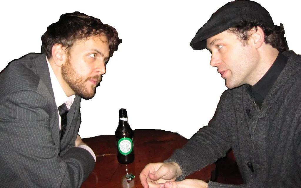 Karl Ward and Kevin Peckham stare intently at each other. A beer is on the table, I wanna say it's an Australian beer? Not a Foster's though, actually maybe it's a Vermont beer? Definitely organic.
