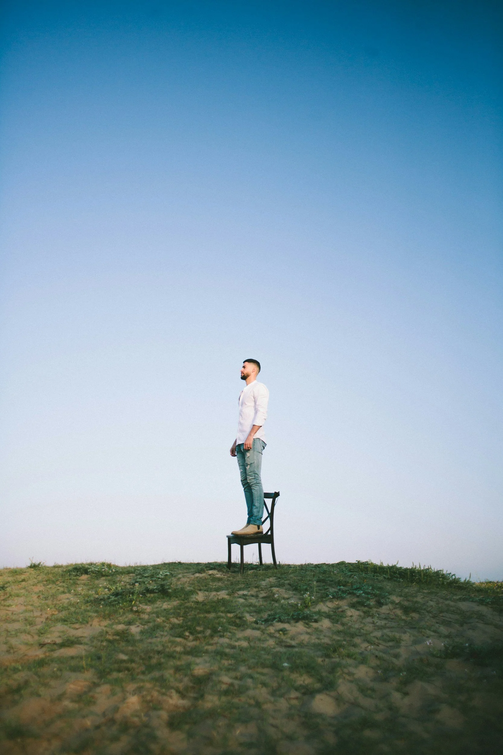 A man stands on a chair in an open outdoor area with a clear sky background.