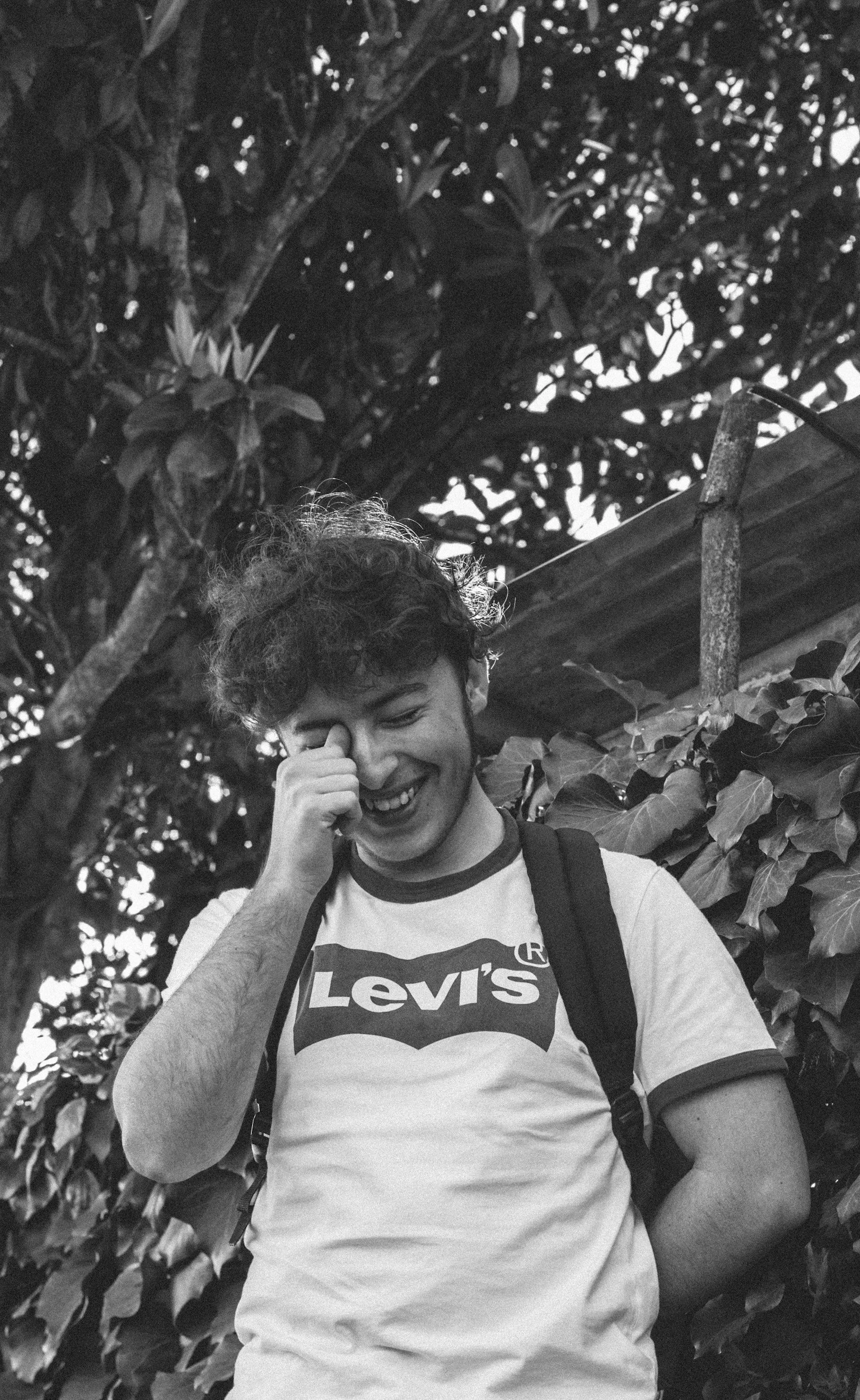 A young man with curly hair is smiling and touching his face with his right hand. He is wearing a white Levi's t-shirt and a backpack, standing outdoors near leafy plants and trees.