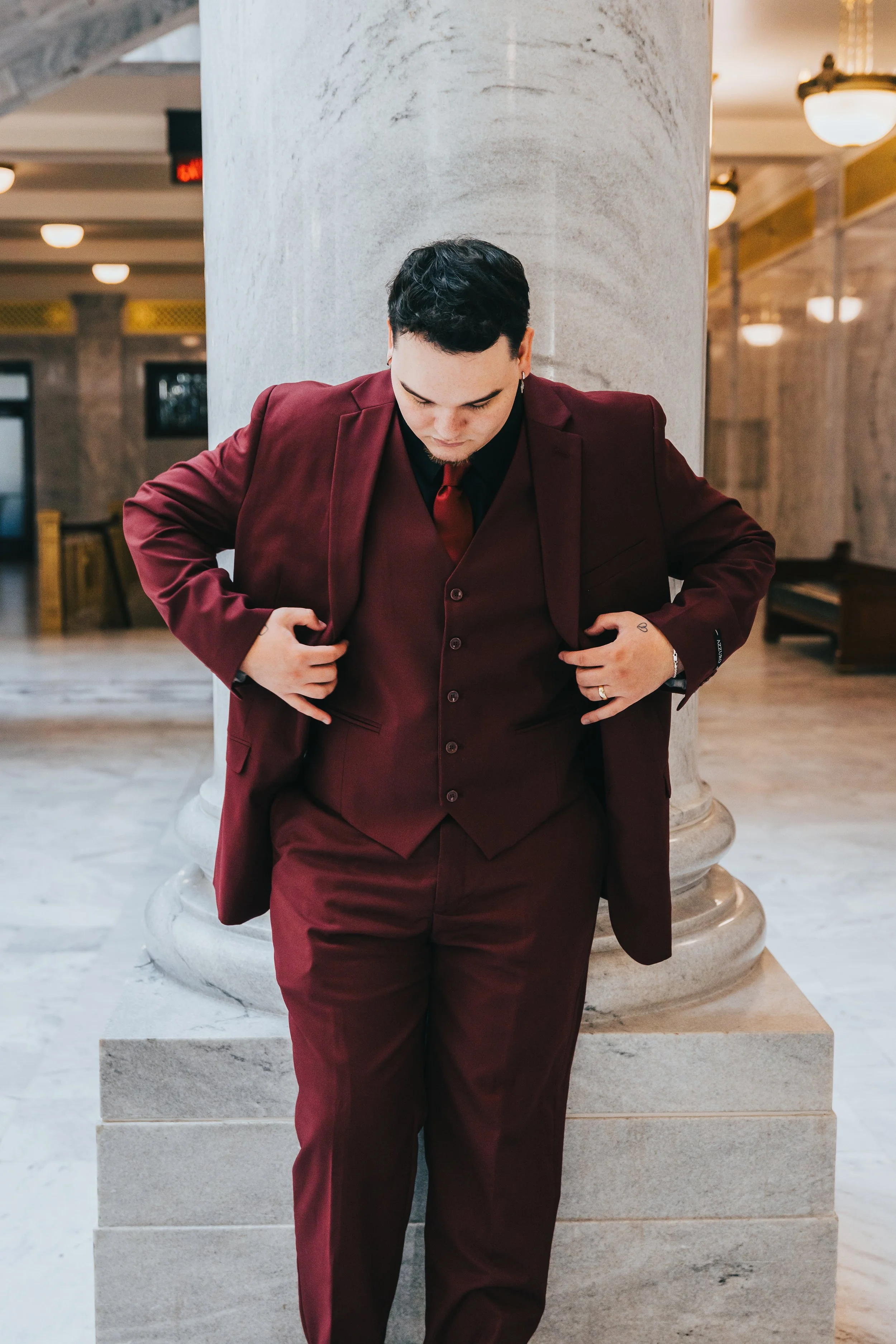 A man in a burgundy suit standing with his head bowed, adjusting his jacket in a marble lobby.