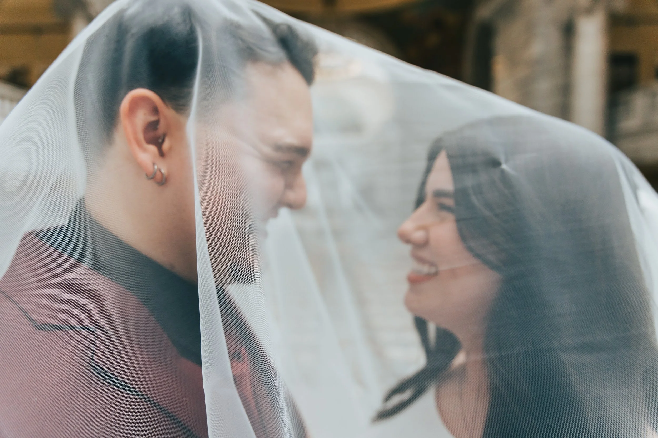 A couple faces each other under a wedding veil, smiling with happy expressions.