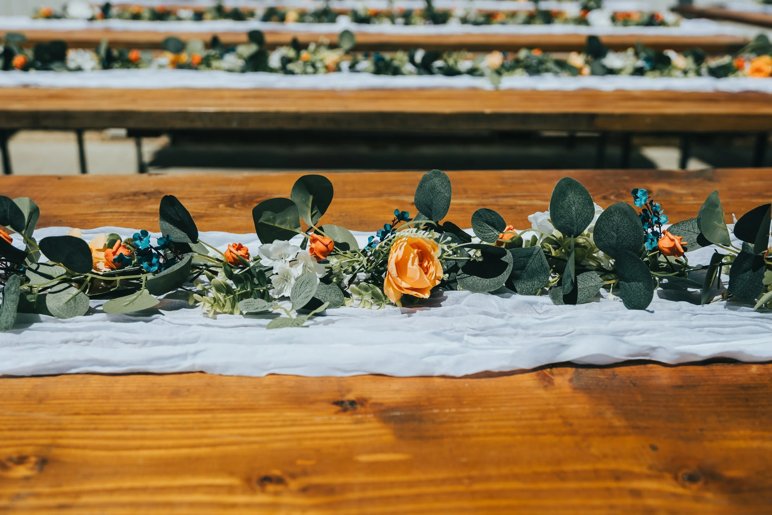 Close-up of a floral arrangement on a white cloth covering a wooden table, with more floral arrangements visible on additional tables in the background, set outdoors.