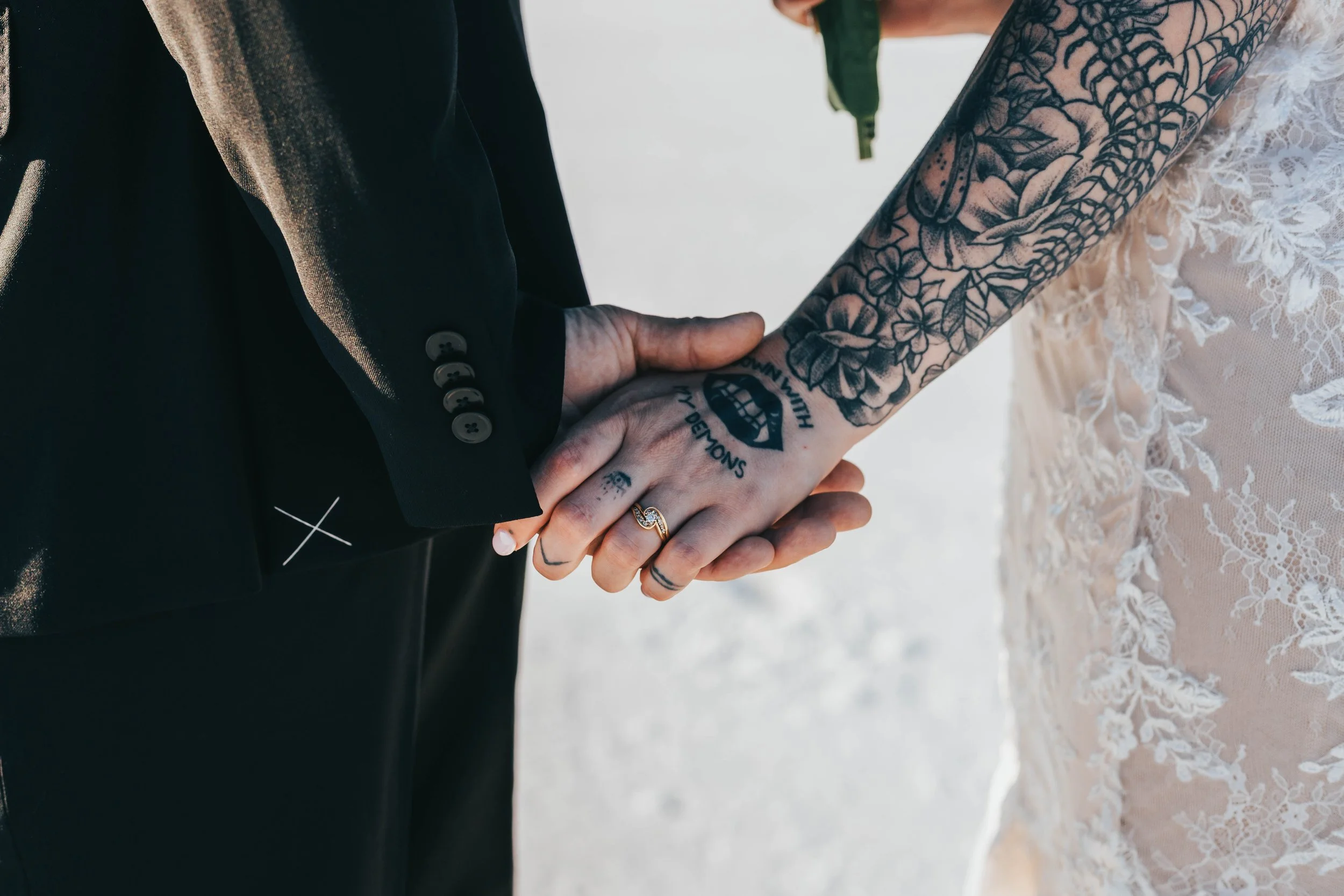 Close-up of a couple holding hands during a wedding, showing tattoos and wedding rings, with the bride wearing a lace dress and the groom in a black suit.