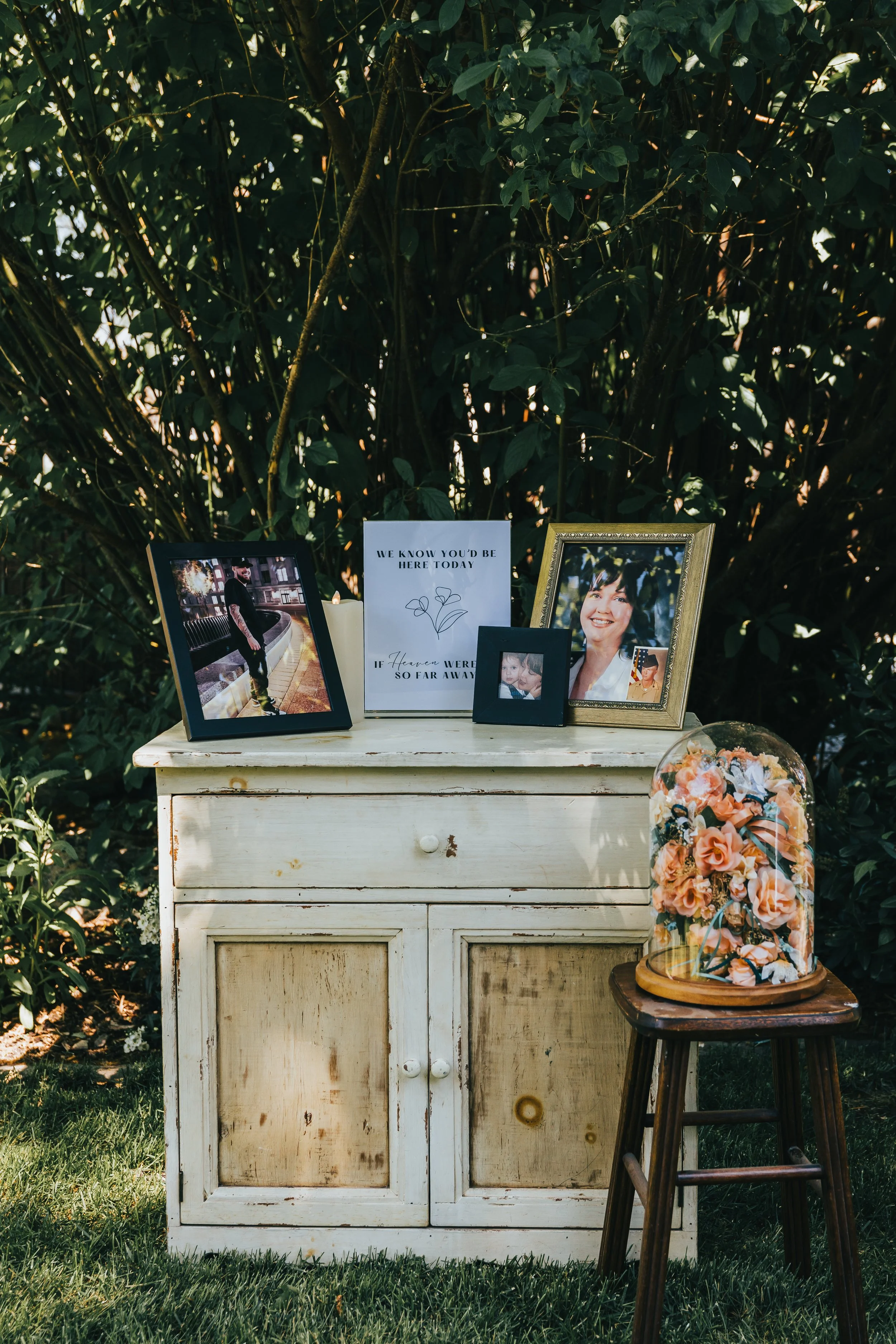 A small, weathered white wooden cabinet on a grassy lawn, topped with framed photographs, a candle, and a sign, with a glass dome holding pink roses on a wooden stool next to it, all set against lush green foliage.