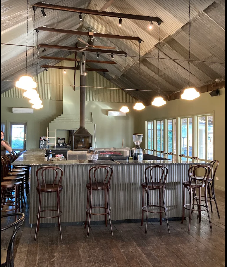 Interior of a cafe or restaurant with a central bar made of corrugated metal, surrounded by high chairs, with a fireplace, large windows, hanging lights, and a decorative ceiling structure resembling a tree or plant.