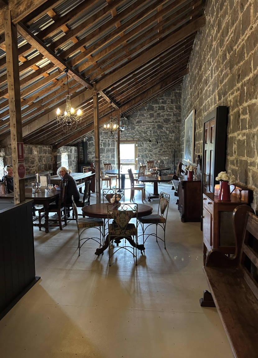 Interior of a rustic restaurant with stone walls, wooden ceiling beams, chandeliers, and various seating arrangements, including tables and chairs, with a couple of people sitting at a table.