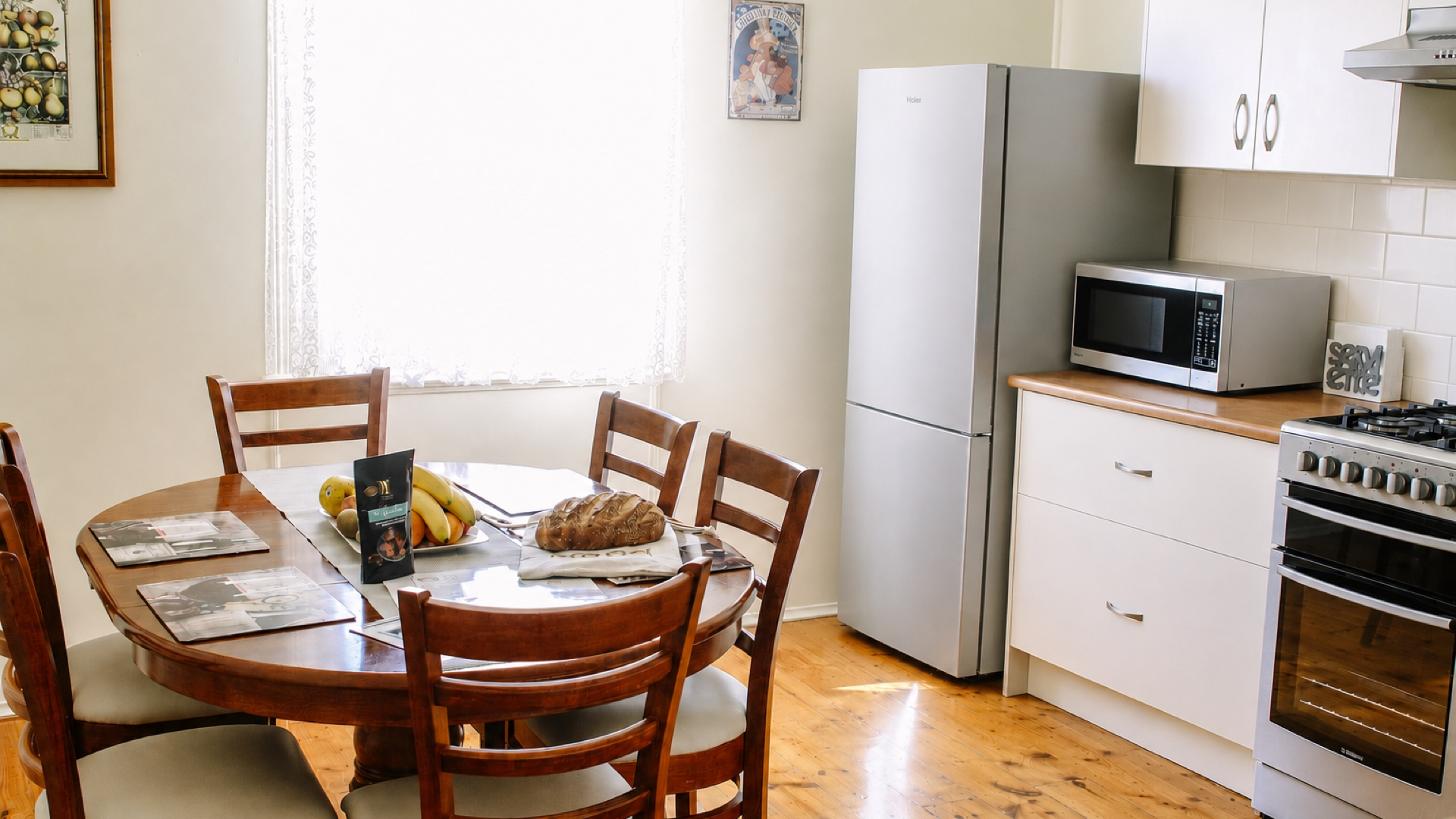 A cozy kitchen with a wooden dining table surrounded by six chairs, a loaf of bread, a banana bunch, and magazines on the table. In the background, there's a white refrigerator, a microwave oven on a wooden countertop, a stove, white cabinets, and a 