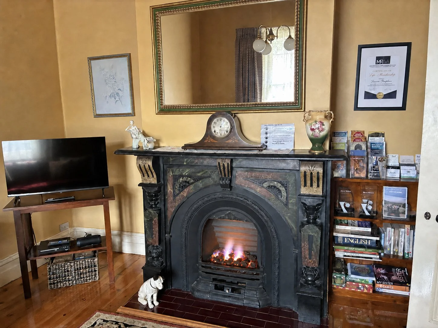 A living room corner featuring a black ornate fireplace with a small fire, a vintage clock on the mantel, a ceramic dog statue, a floral vase, a mirror above the fireplace, a flat-screen TV on a wooden stand, a wooden bookshelf filled with books and 