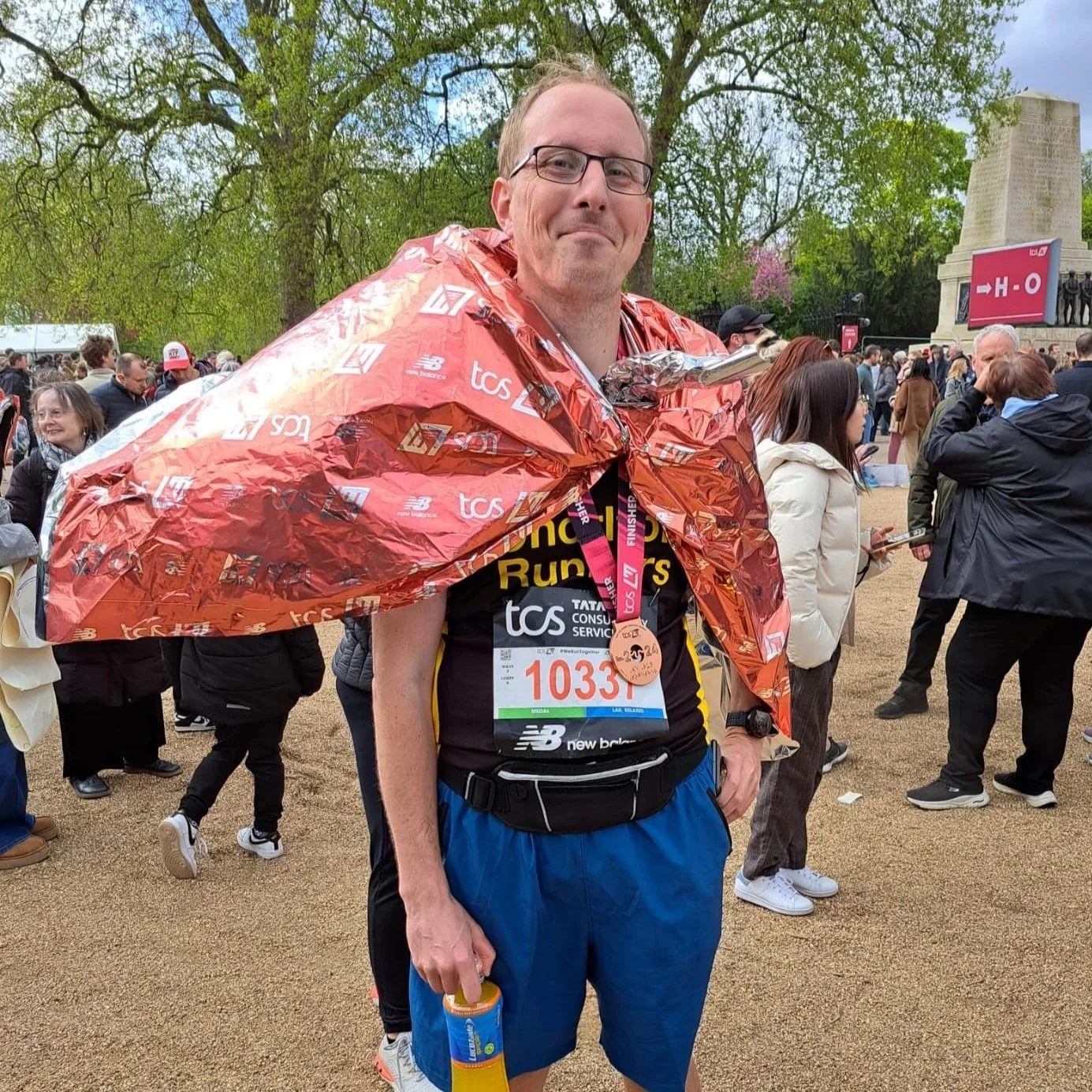 A man in running clothes with a race bib, wearing a medal around his neck, and holding a water bottle, standing among a crowd at a race event, with trees and a monument in the background.