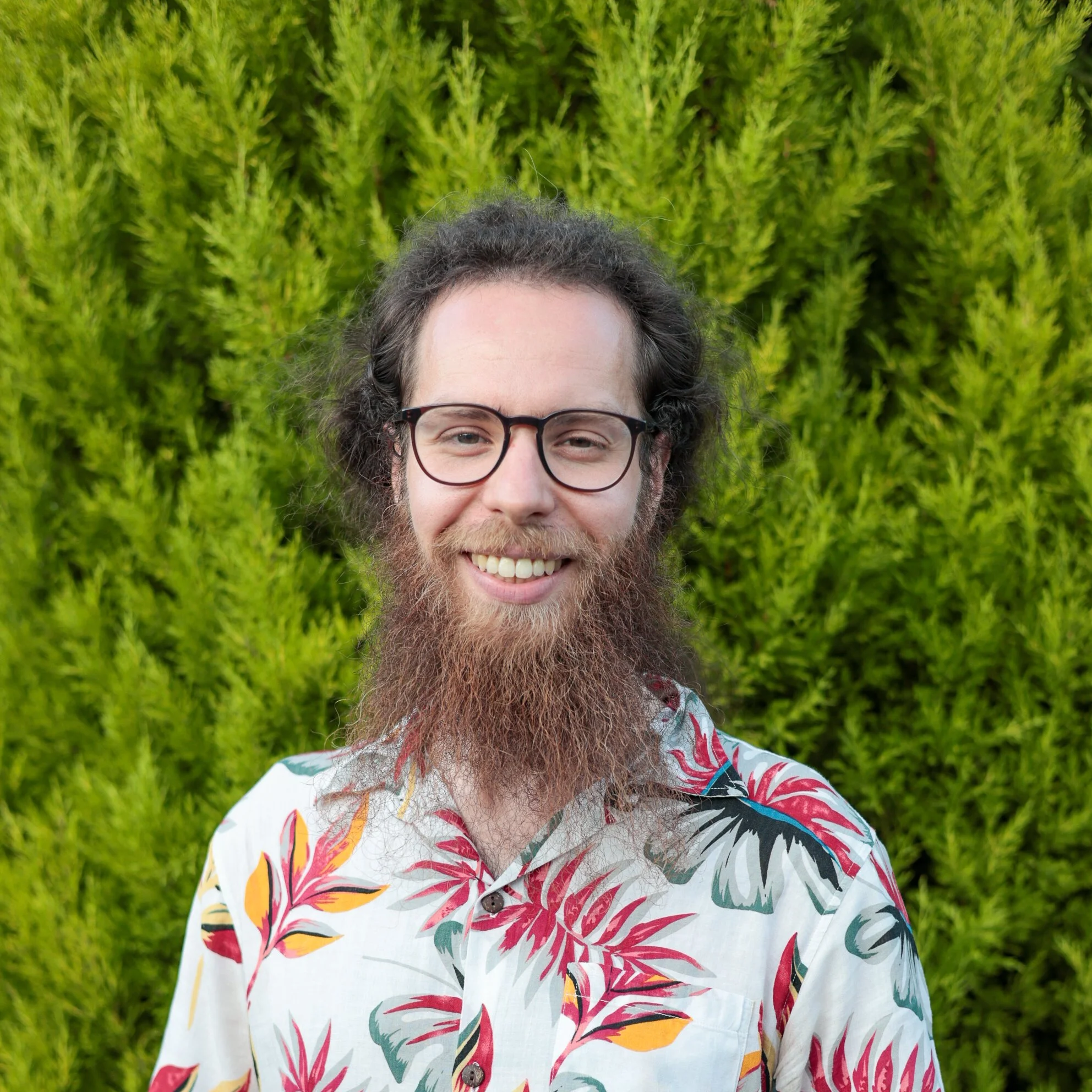 A young man with long, curly brown hair and a thick beard taking a selfie against a plain white background.