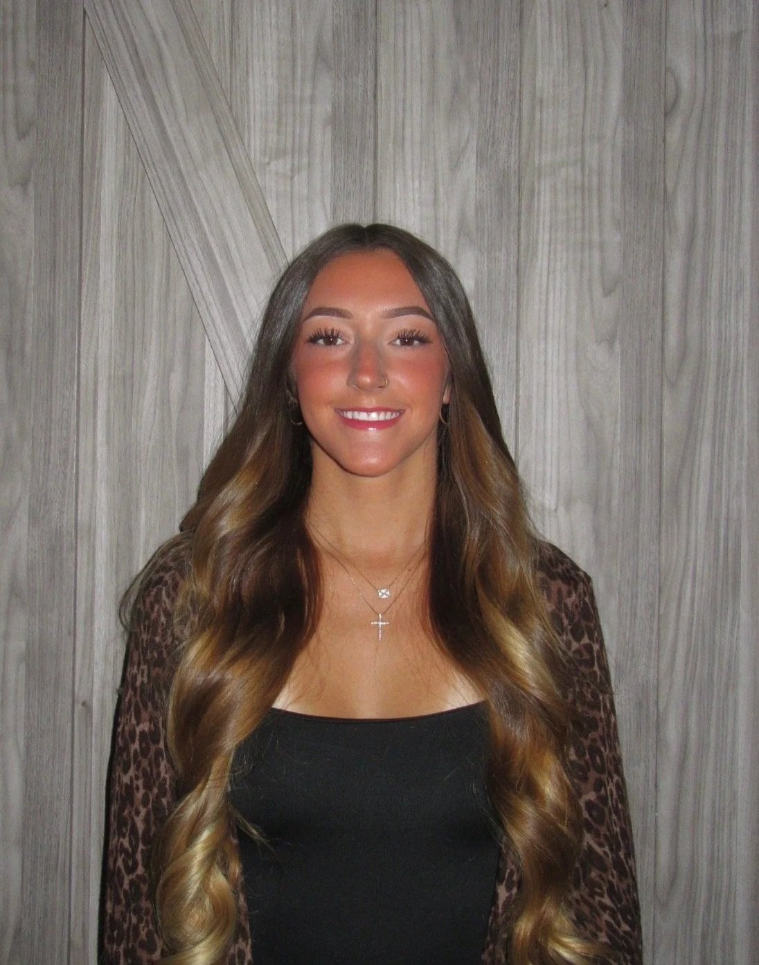 A young woman with long wavy hair, smiling, wearing a black top and a leopard print jacket, standing in front of a wooden background.