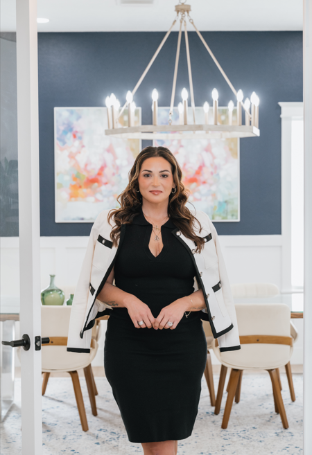 A woman in a black dress and white jacket stands in a modern dining room with a chandelier and colorful abstract painting behind her.