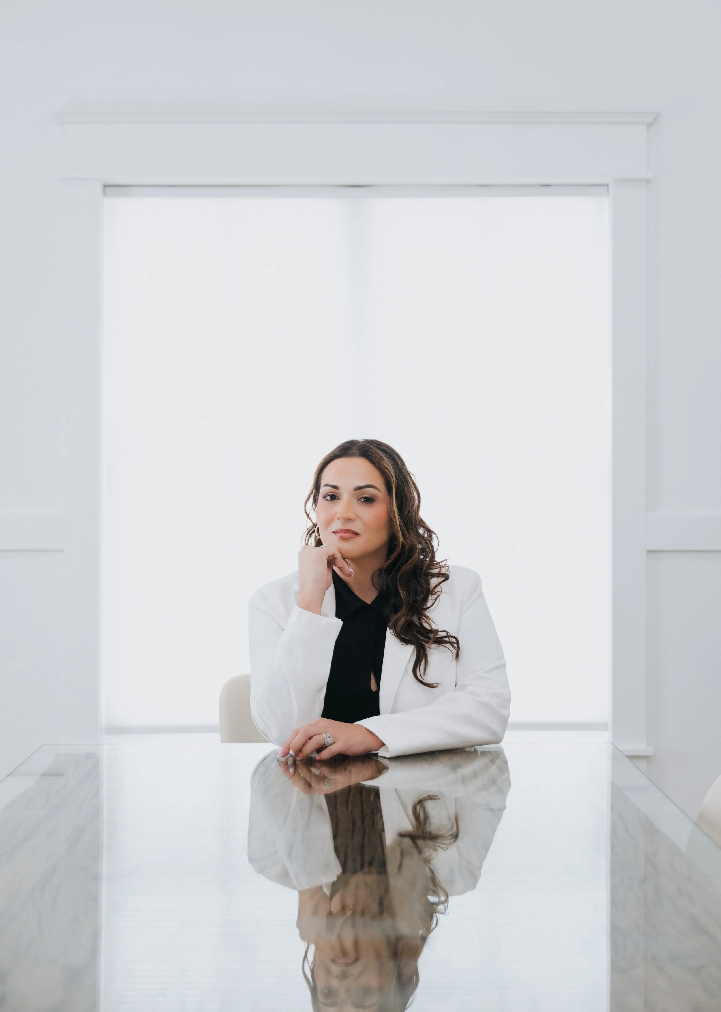 A woman with long, wavy brown hair, wearing a white blazer over a black top, is seated at a reflective marble table in a bright, modern room with white walls and a large window behind her.