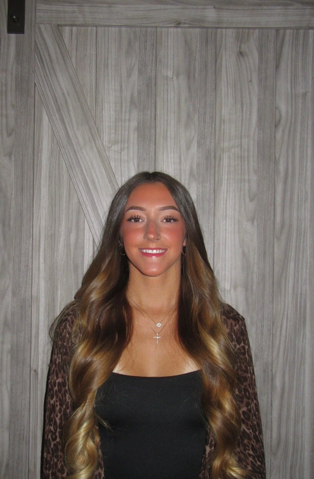 A young woman with long, wavy brown hair smiling, standing in front of a gray wooden door. She is wearing a black top, a leopard print cardigan, and layered necklaces.