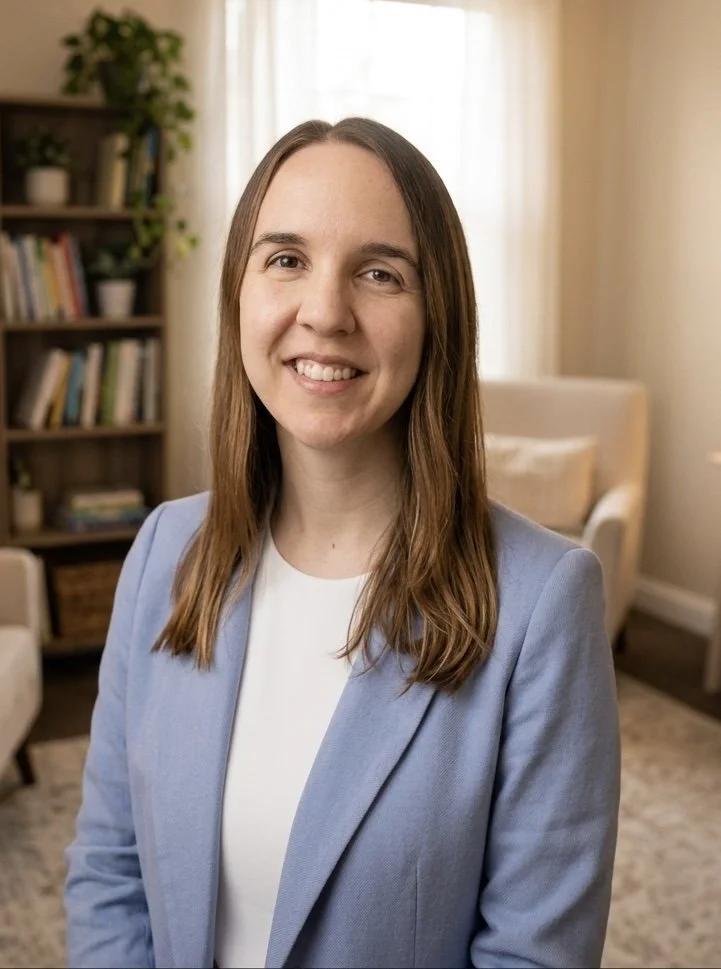 A woman with long brown hair and a light blue blazer smiling in a cozy living room with bookshelves, a beige armchair, and a window with curtains.