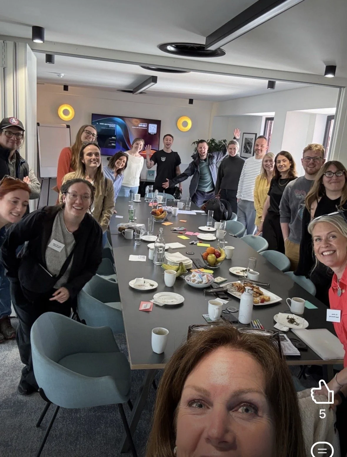 Group of people gathered around a conference table in a modern meeting room, smiling and posing for a photo. The table has plates with food, cups, bottles, and various notes and items on it.
