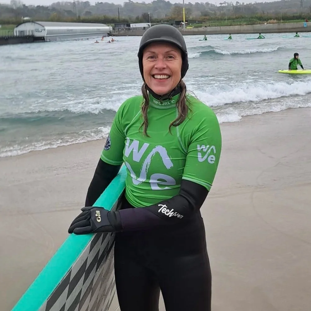 Woman in green wetsuit and black helmet smiling at the beach, holding a paddleboard, with others surfing and paddleboarding in the background.