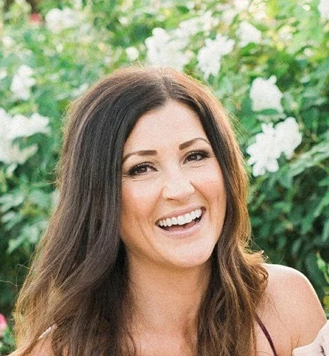 A holistic therapist in Encinitas with long brown hair smiling outdoors with white flowers and green foliage in the background.