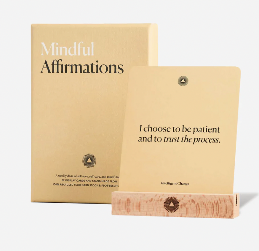 A box labeled 'Mindful Affirmations' positioned behind a card that reads, 'I choose to be patient and to trust the process,' with a small logo at the bottom of the card and the box, and a pink stand holding the card.