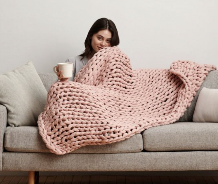Woman sitting on a beige sofa, holding a mug, covered with a large pink chunky knit blanket.