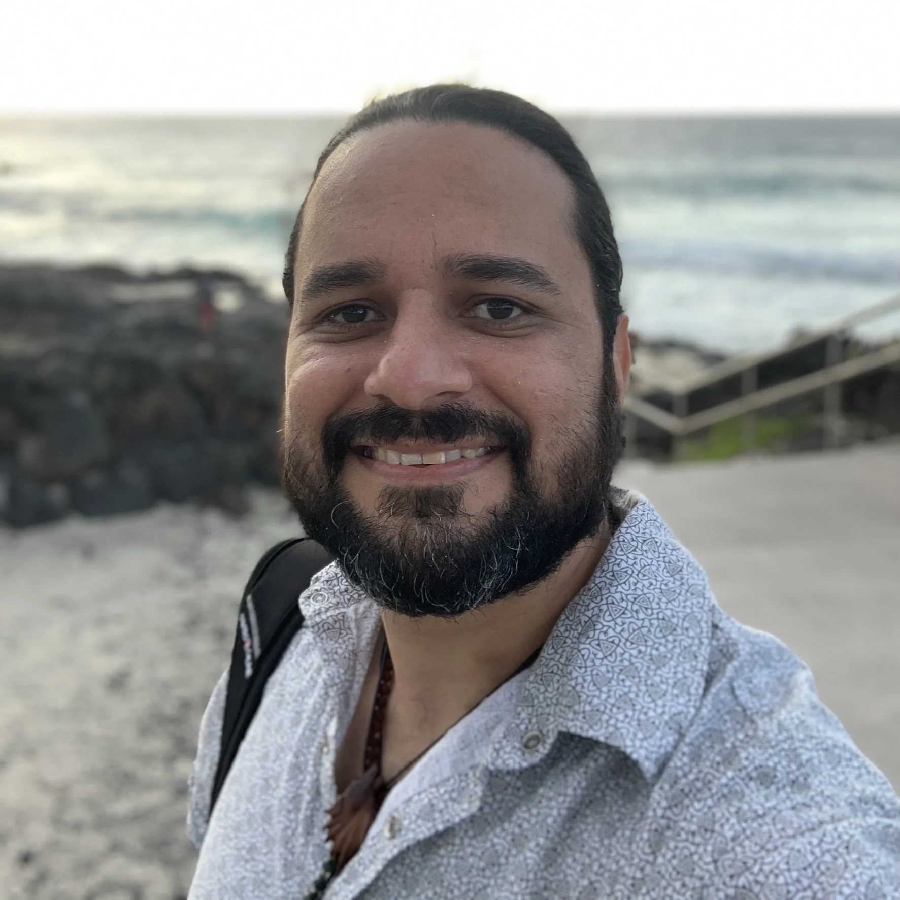 Sergio Saenz, medicine musician & cofounder of Agape Church wearing a light patterned shirt, taking a selfie at the beach with ocean waves and rocks in the background.