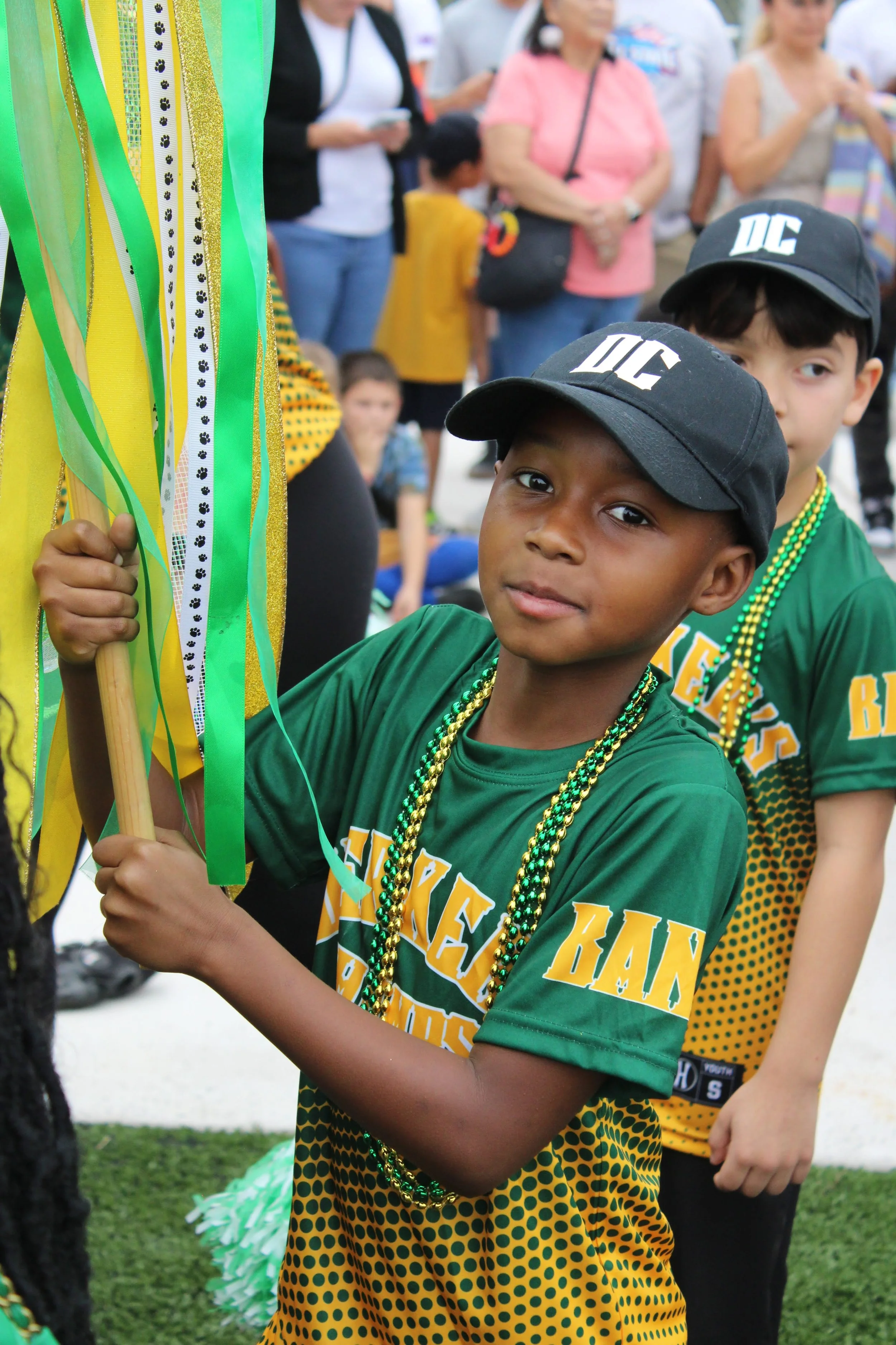 A young boy holding green and yellow streamers during a festive parade or celebration. He is wearing a green shirt with yellow text, green and gold beads, and a black cap with white letters.