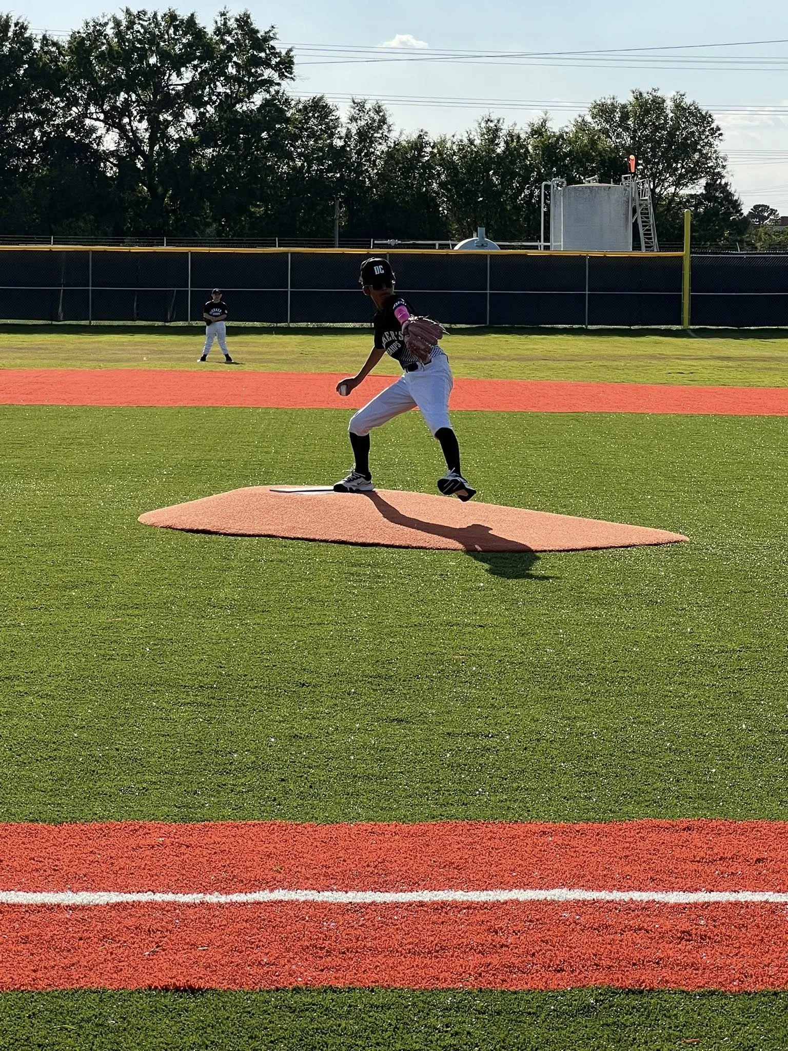 A young baseball player standing on the pitcher's mound, preparing to pitch during a game on a green field with red foul lines, with another player in the background and trees in the distance.