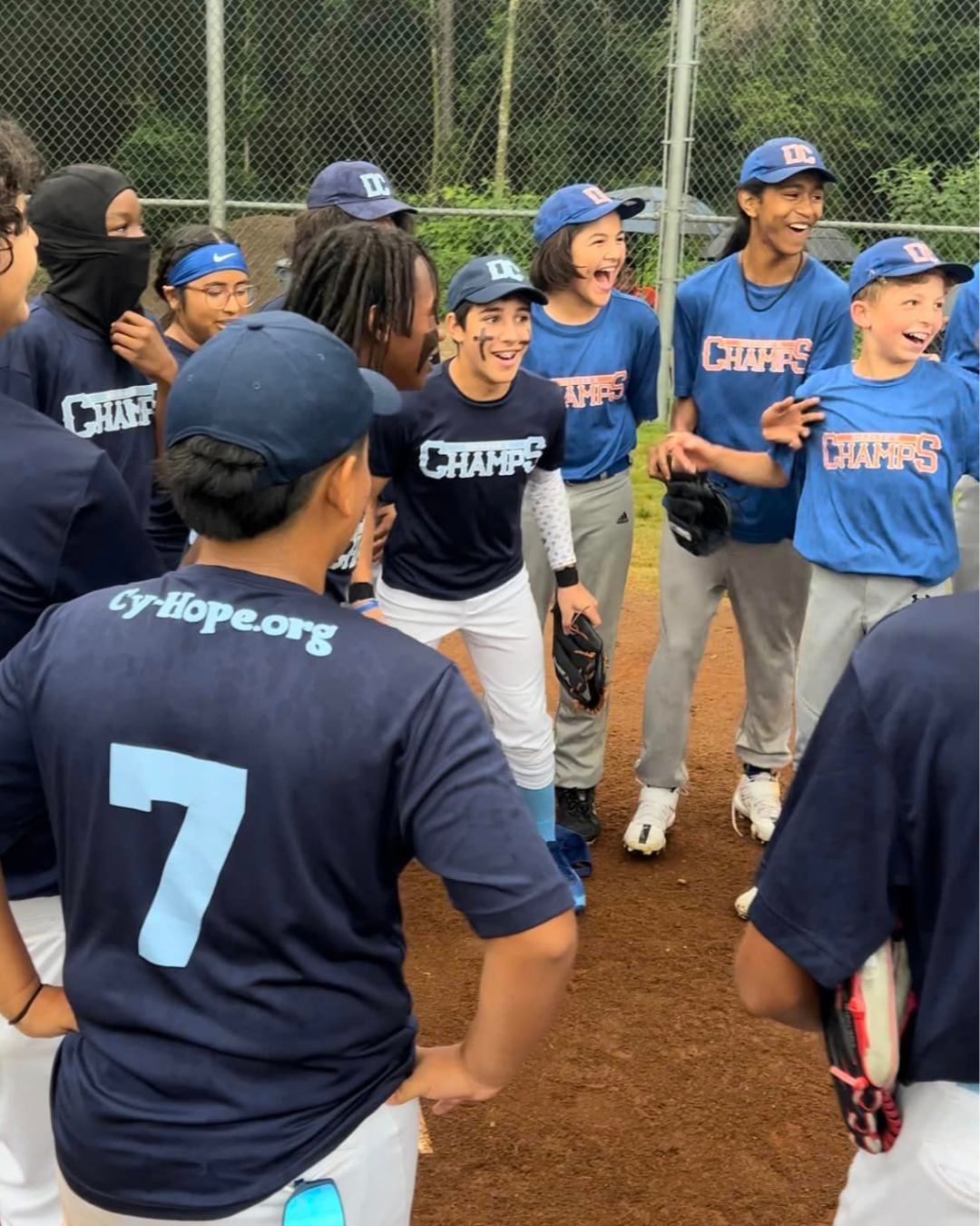 A group of young softball players in blue and black jerseys with the word 'CHAMPS' on them, standing on a dirt field and laughing together after a game.