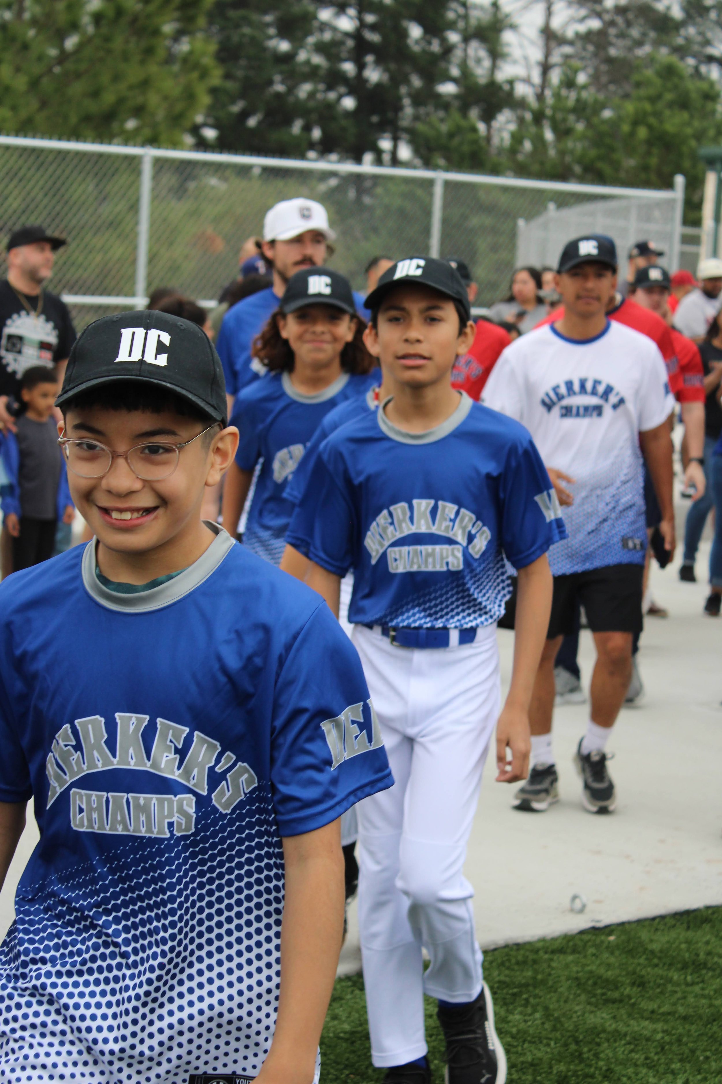 Group of children and adults in baseball uniforms and caps walking outdoors on a cloudy day, with a chain-link fence and trees in the background.
