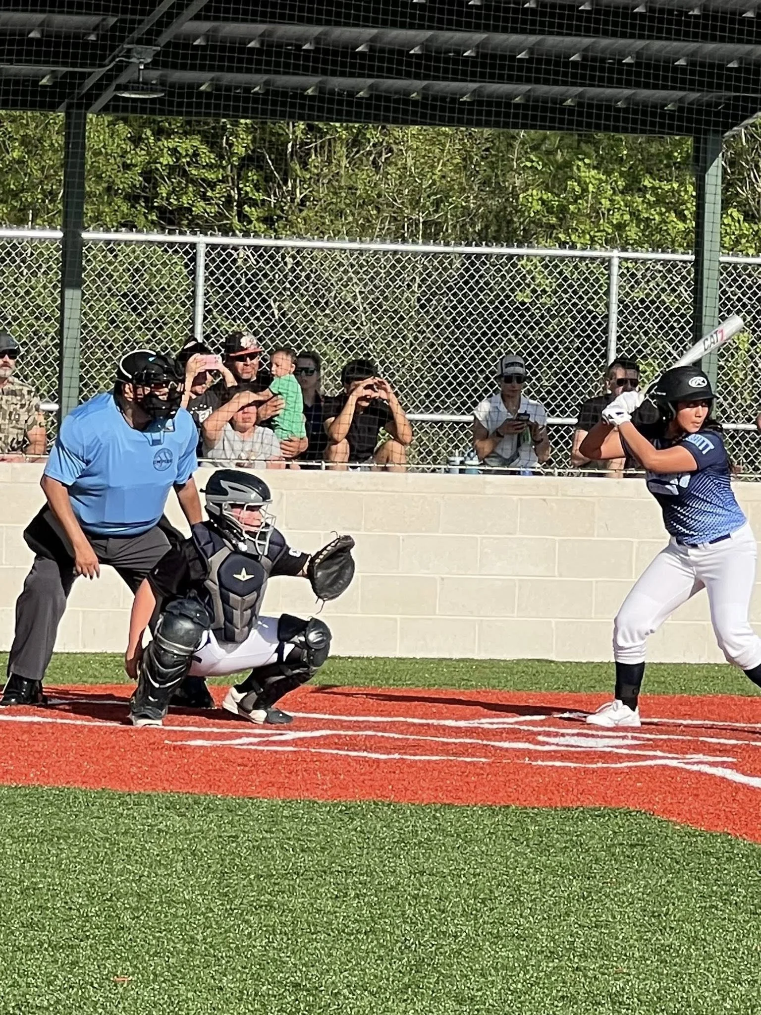 A female softball batter at the plate ready to swing, a catcher crouched behind her, and an umpire in blue behind the catcher, with spectators watching behind a chain-link fence.