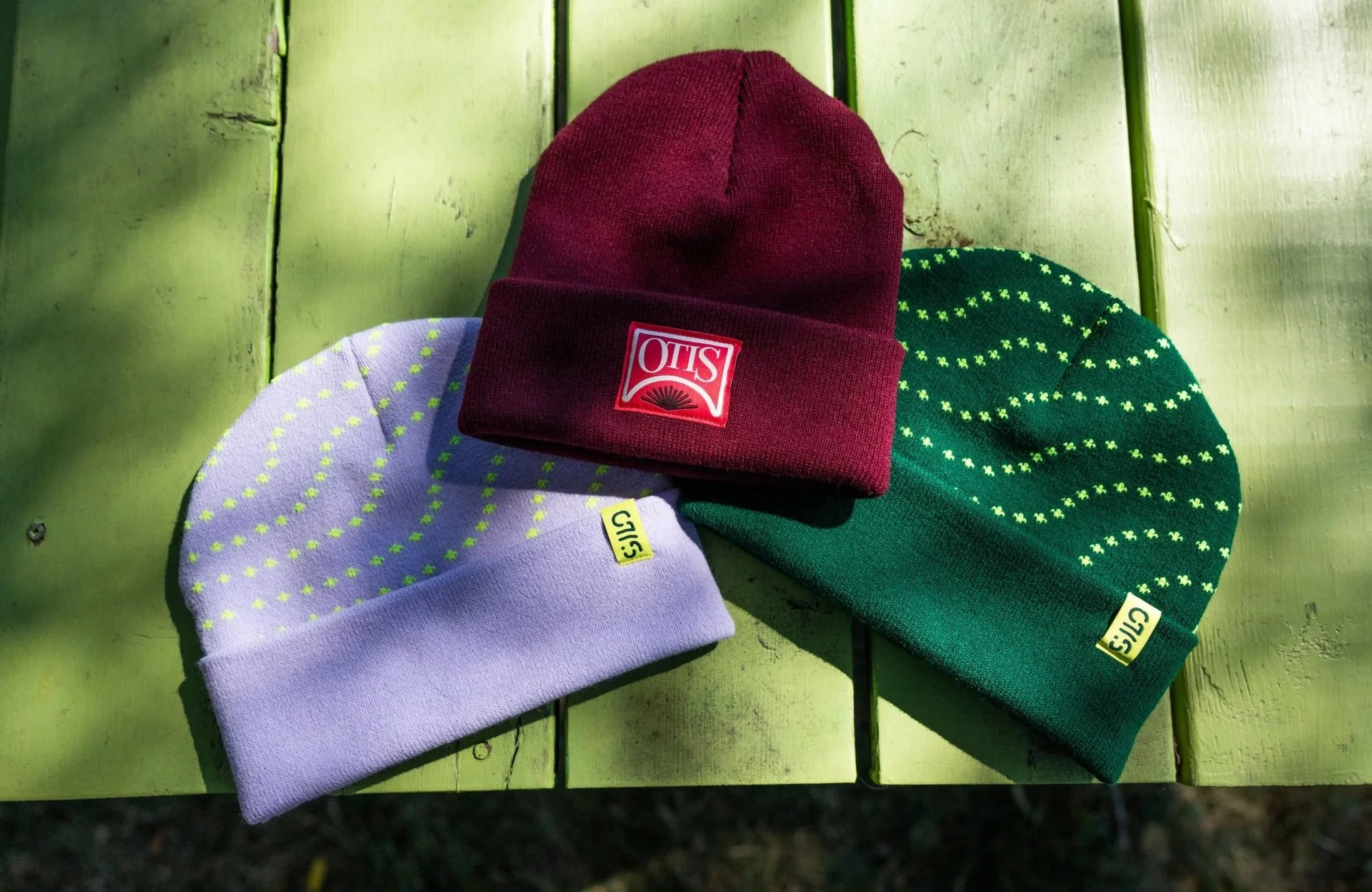 Three knit Otis Mountain Get Down beanies in red, purple, and green on a green wooden surface.