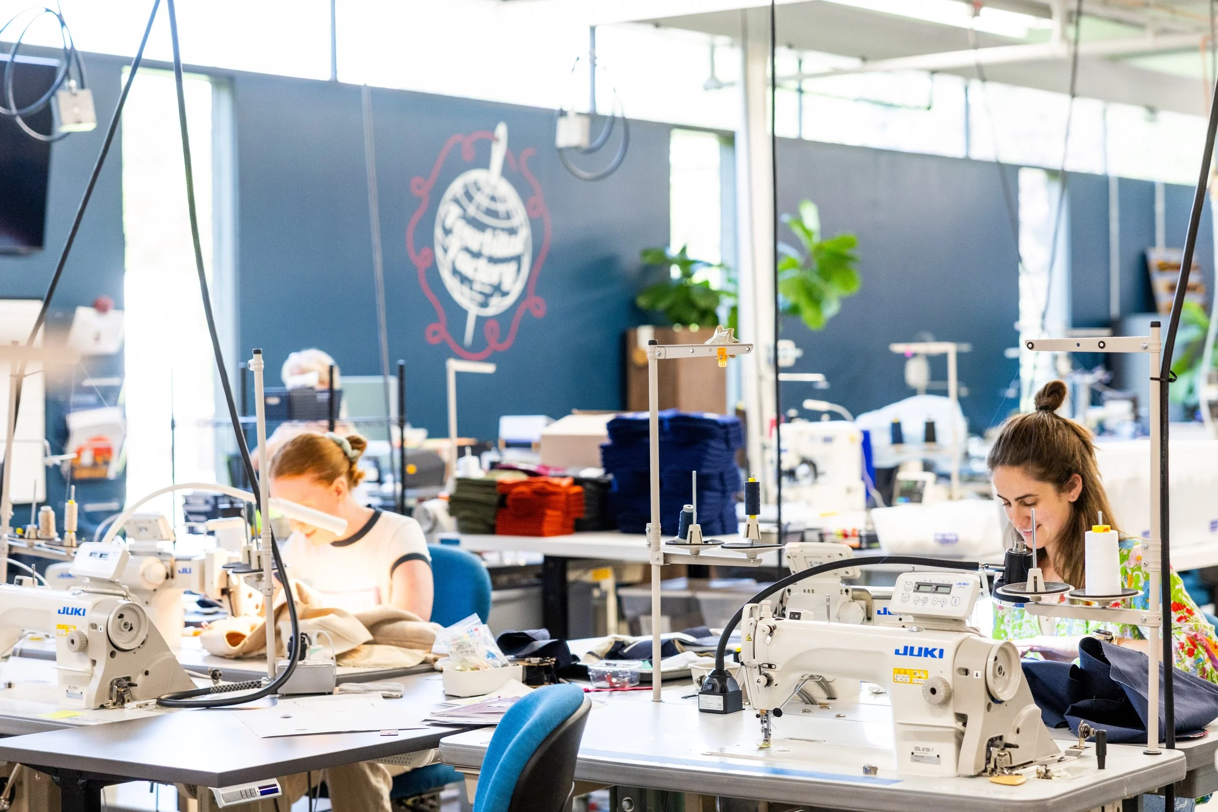 Two women working at sewing machines in a textile workshop with fabric and supplies, colorful fabrics stacked on tables, and a decorative wall art of a blueberry on a dark blue wall.