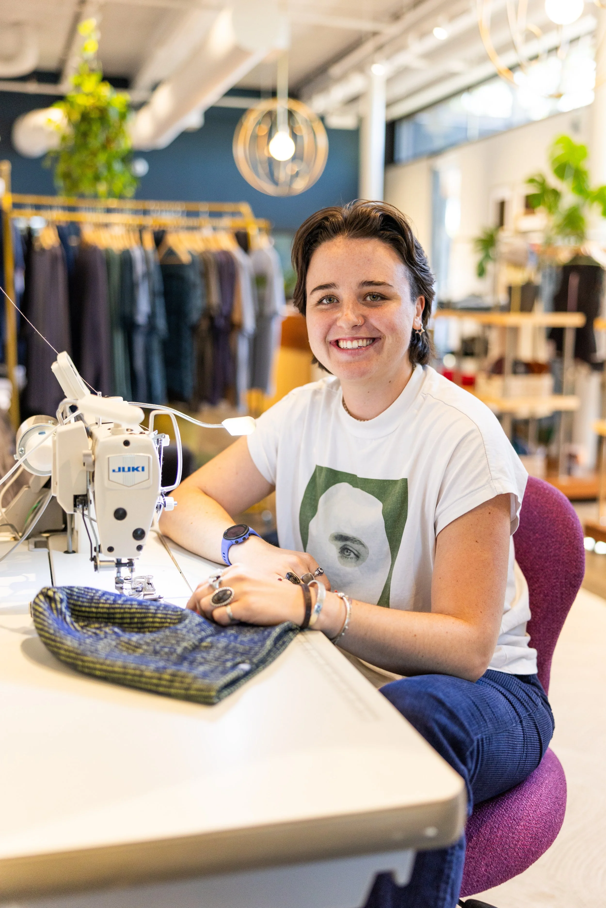 Young woman sitting at a sewing machine in a clothing boutique or workshop, smiling at the camera.