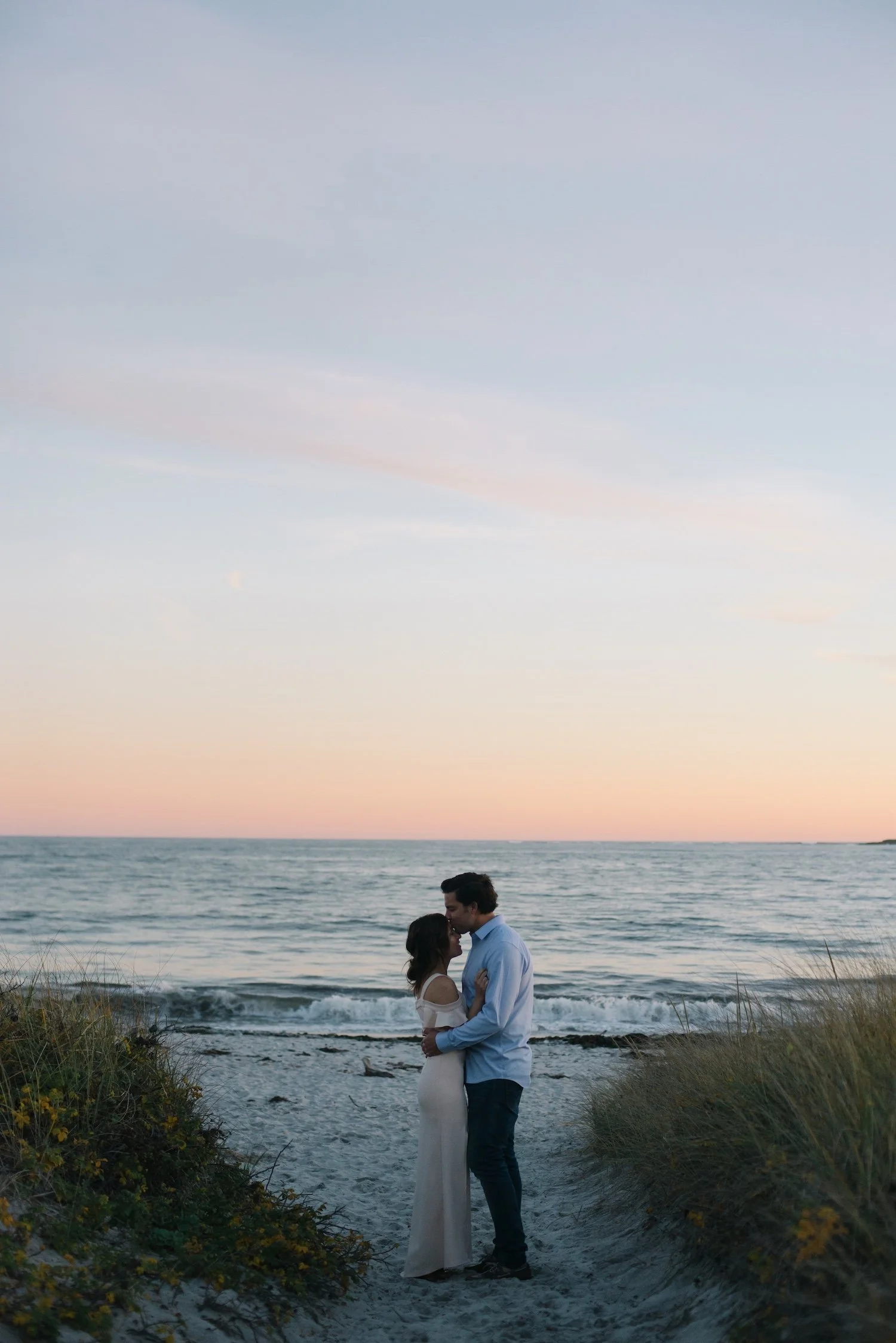 Portrait session on the beach in Maine. Couple embracing on the beach with waves behind them at twilight.