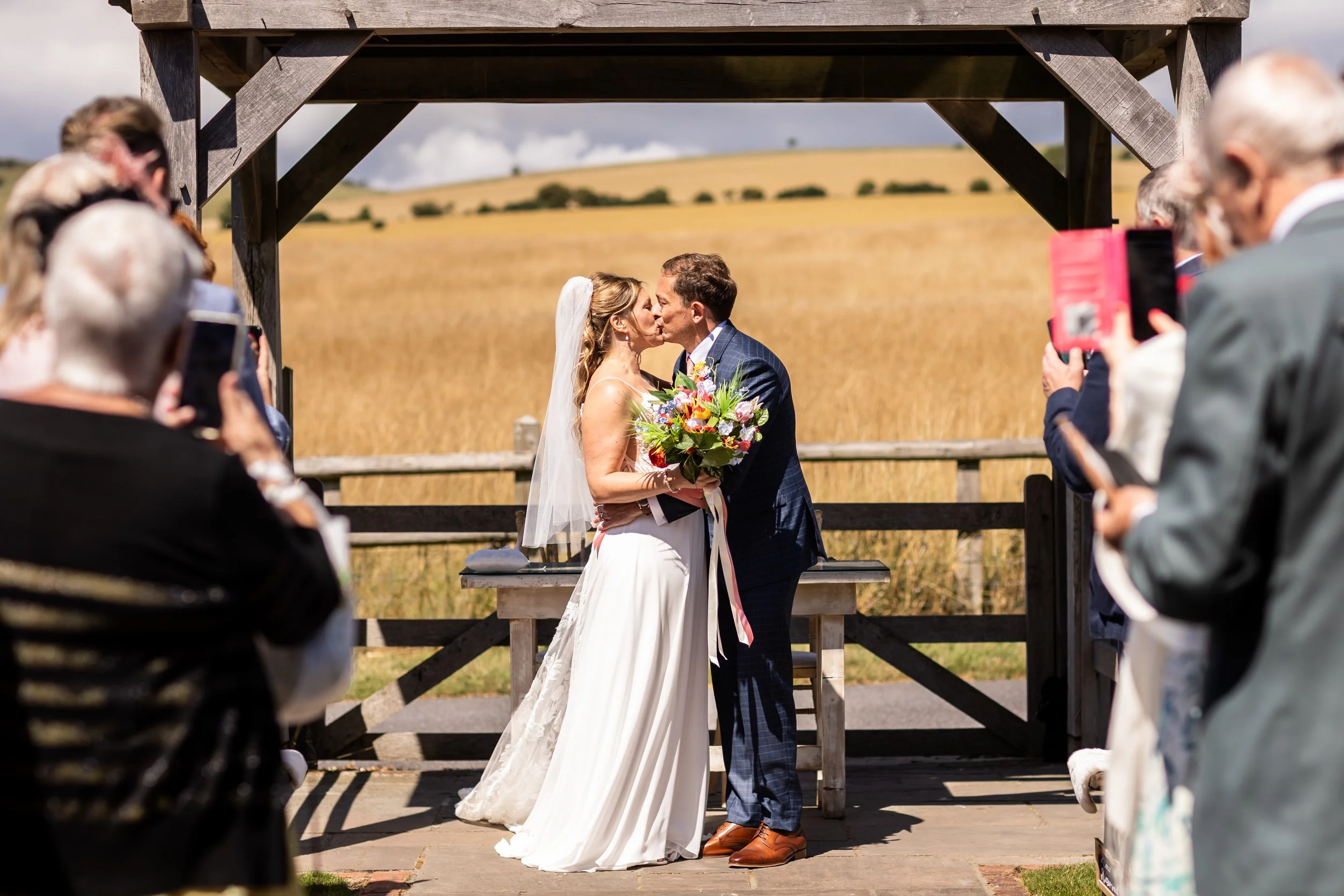 Anna and Christian sharing their first kiss as newlyweds during their Long Furlong Barn wedding ceremony surrounded by colourful silk wedding flowers.