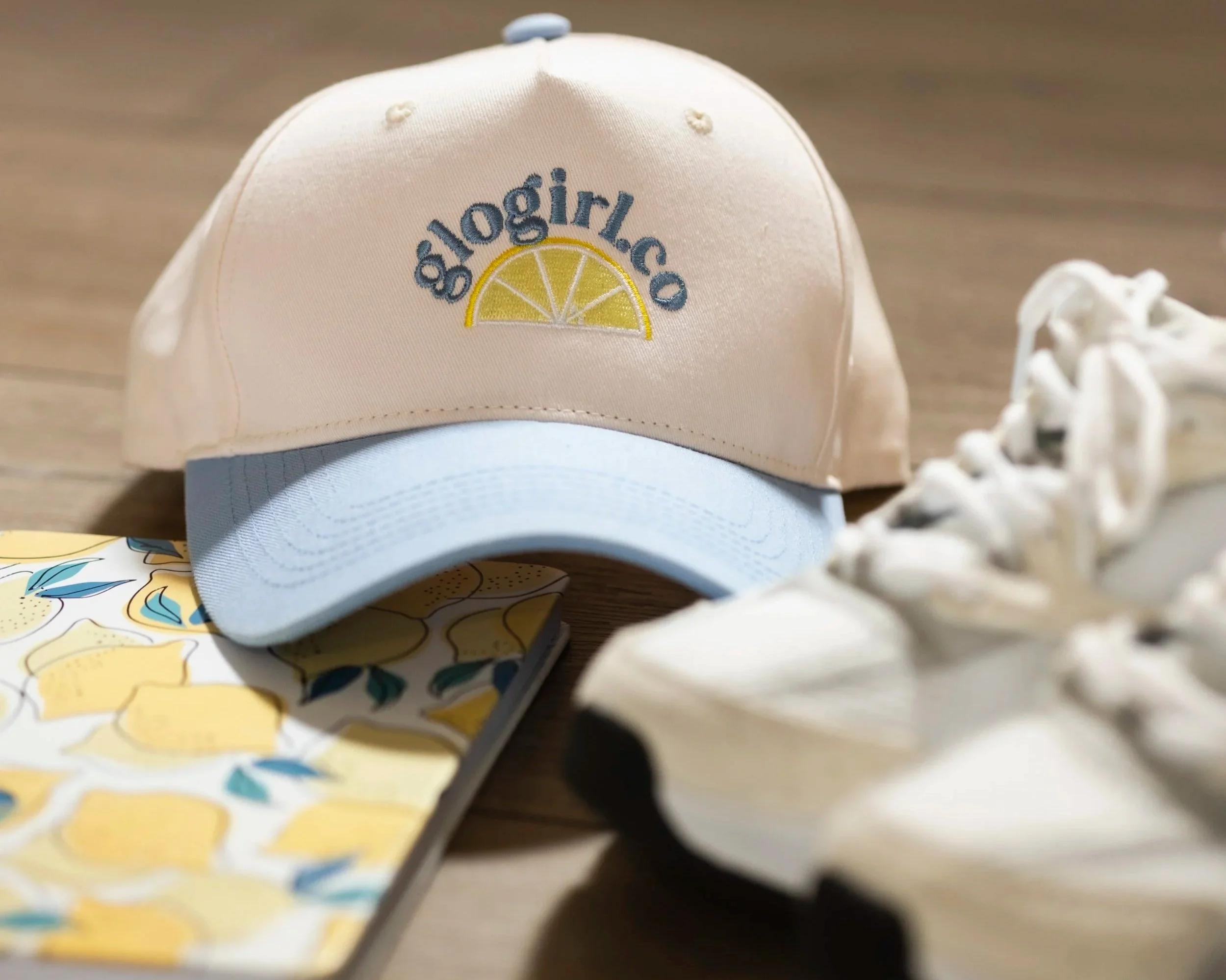 A pastel-colored baseball cap with a lemon slice and the text 'sglogirloco' embroidered on it, next to a notebook with lemon illustrations, and a pair of white sneakers, all on a wooden surface.