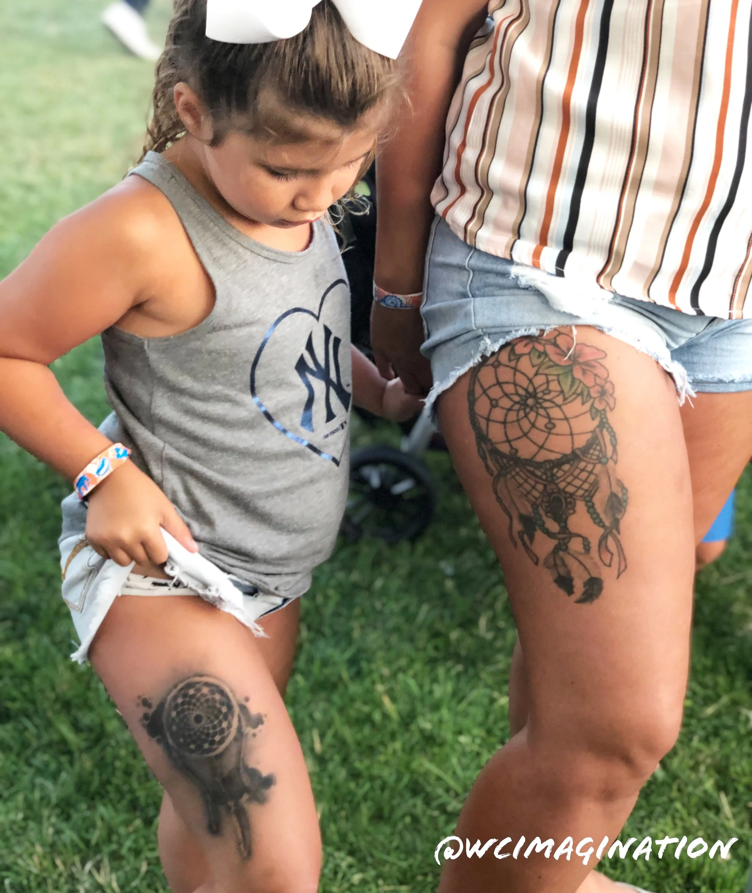 Child and adult with matching dreamcatcher tattoos on thighs, wearing casual summer clothes, standing outdoors on grass.