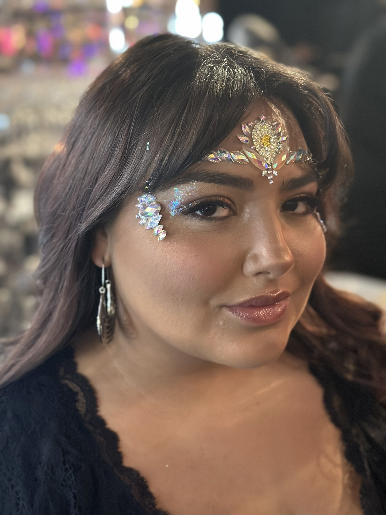 Woman wearing rhinestone face decorations and earrings, with long dark hair, in a softly lit setting.