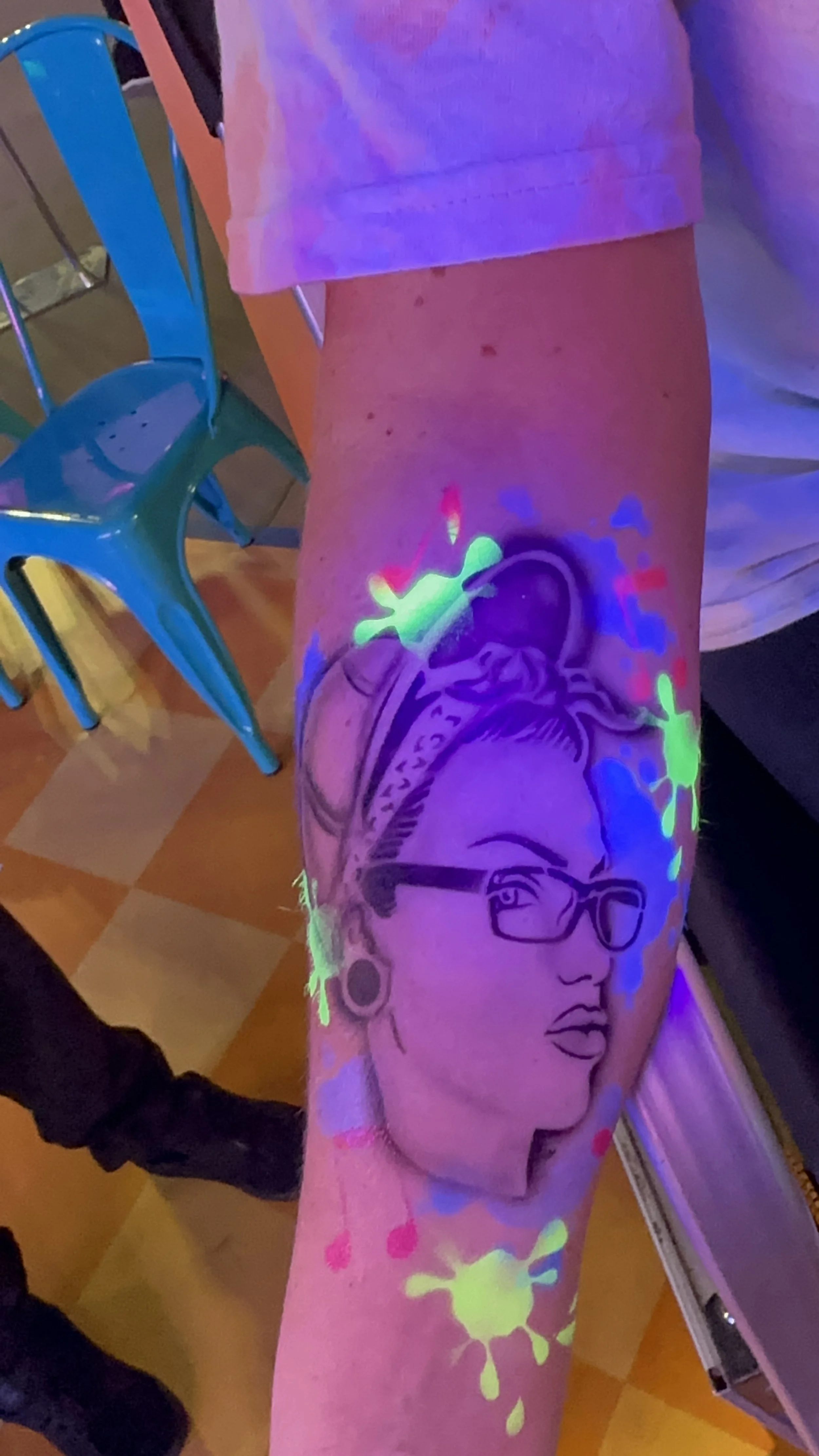 UV tattoo of a woman wearing glasses and bandana on forearm, with colorful neon splatters.