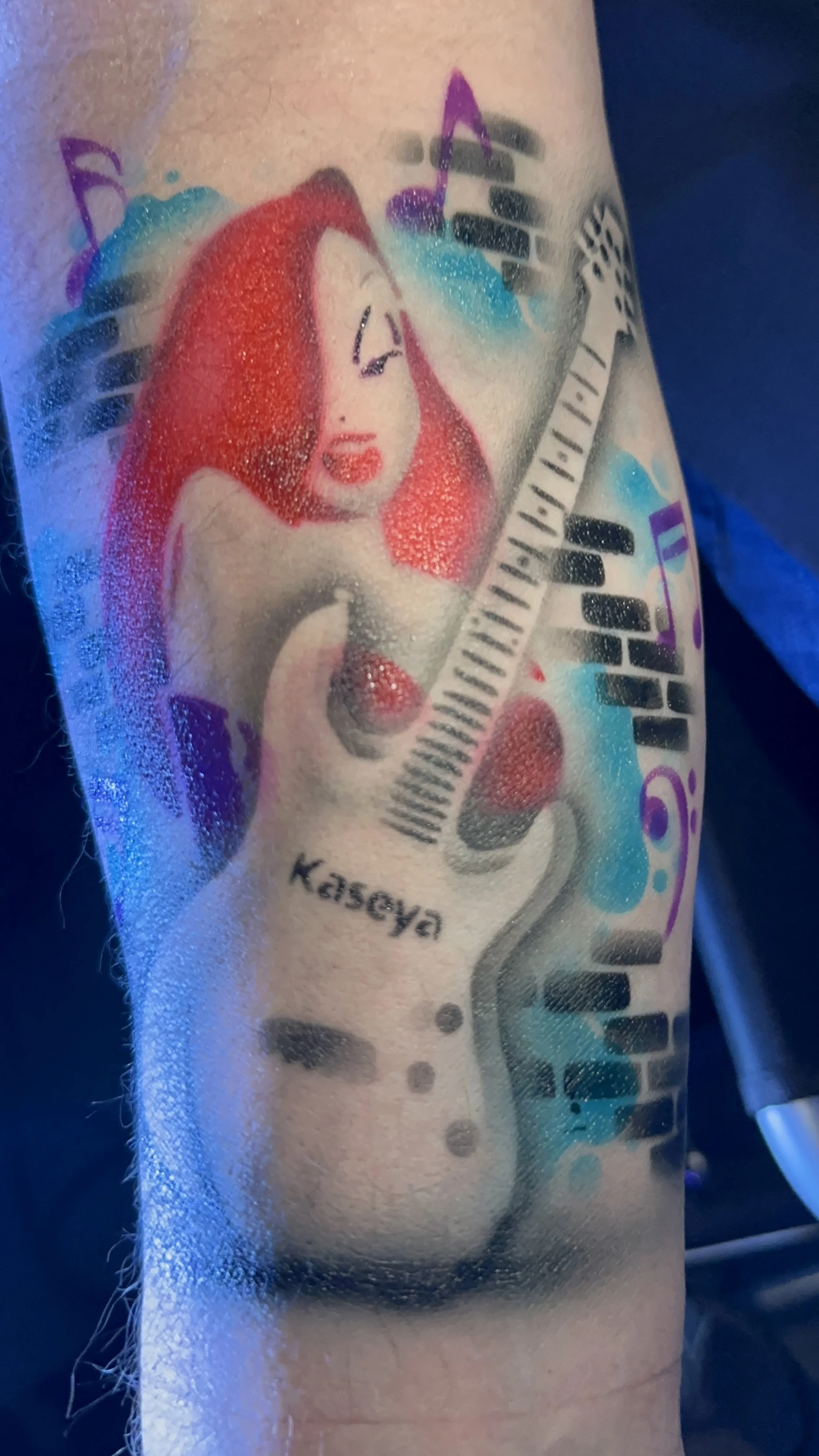 Tattoo of a character with long red hair holding a guitar, colorful musical notes, and the name 'Kaseya'.