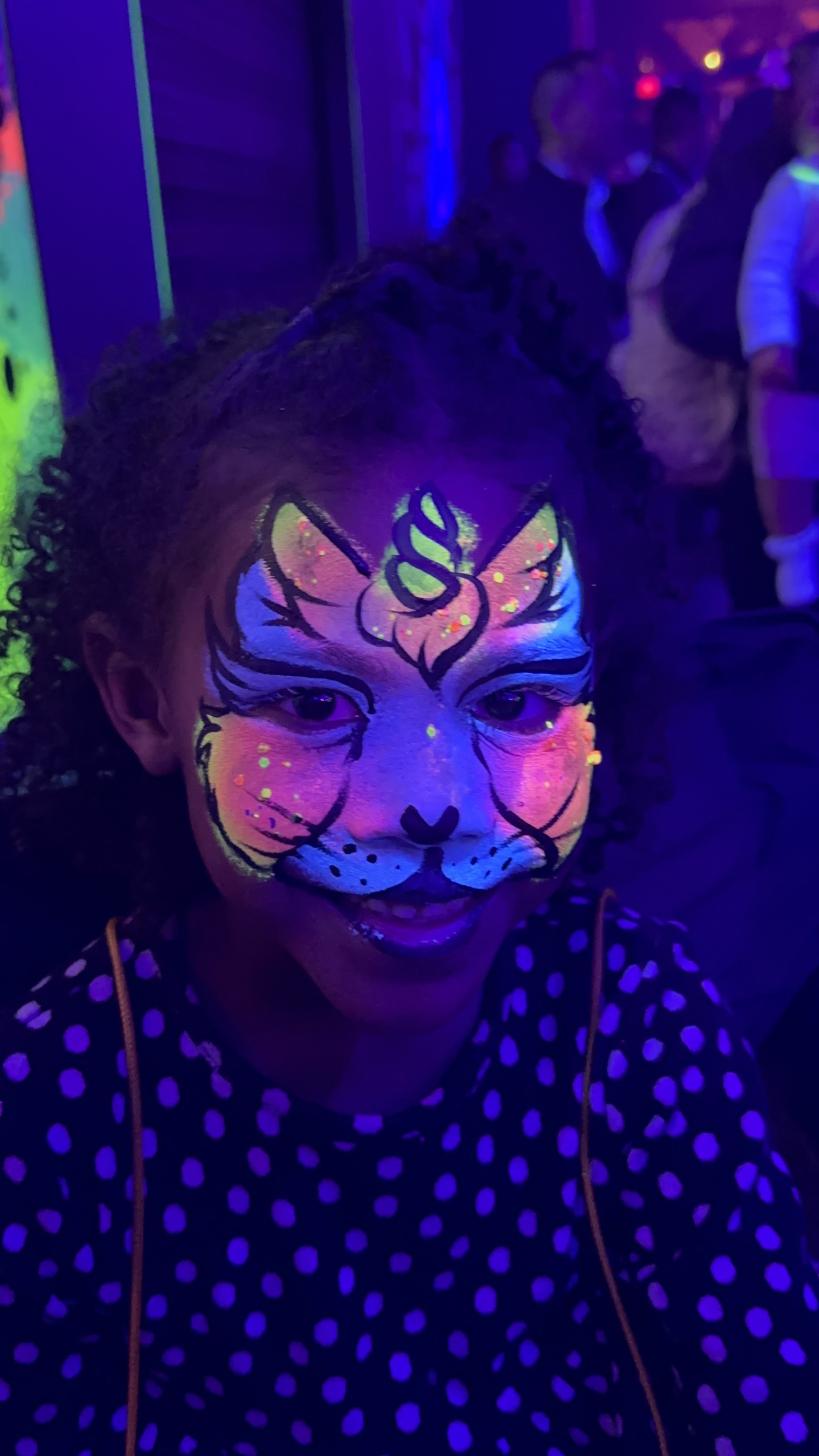 Child with neon face paint resembling a cat under blacklight, polka dot shirt, party setting.
