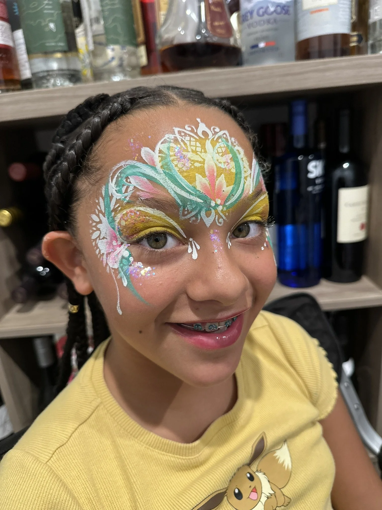 Young girl with braided hair and colorful face paint, wearing a yellow shirt with a cartoon character, smiling in front of a shelf with bottles.